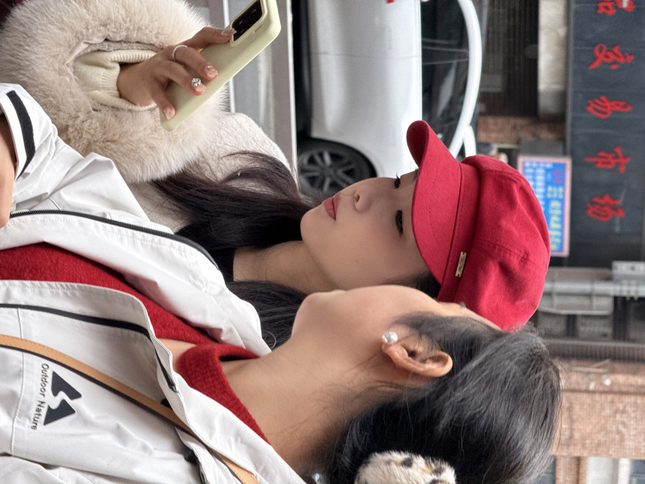 The image depicts two women in an urban street scene within Yuzhong District, China. The woman positioned on the left, slightly behind and to the left of the other, wears a solid red newsboy-style hat with a small gold-colored rectangular emblem above the brim. She has long dark hair and is dressed in a light-colored, possibly cream or beige, fur-trimmed coat. Her gaze is directed downwards at a light green smartphone she holds vertically in her right hand. Her right hand displays manicured nails and a silver-colored ring with a clear stone on her ring finger.
The second woman, whose right profile and upper torso are visible in the foreground on the right, wears a white jacket with a black trim and an "Outdoor Nature" logo on the right chest. Underneath, a textured red garment with a pointed collar is visible. Her dark hair is pulled back, secured by a leopard print hair accessory, and she wears a round white pearl stud earring in her visible left ear. Her head is turned towards the first woman.
The background shows a blurred urban environment with a partially visible white sedan-style car behind the women. Above the car, a building facade features a dark signboard displaying red Chinese characters, "交易市场" (Jiaoyi Shichang, meaning "Trade Market"), and a smaller blue sign with white text. Metal poles and street infrastructure are also present.