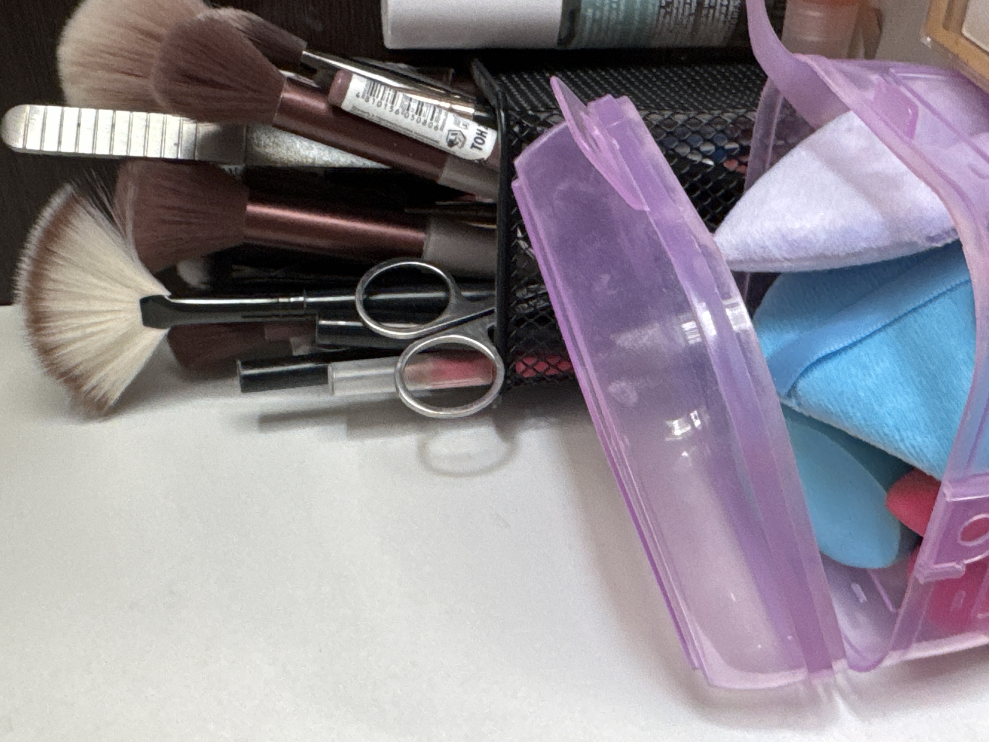 The image shows a close-up of makeup brushes and other beauty products arranged in a container. The items are mostly in shades of brown, pink, and blue, with a few metallic accents. In the foreground, a clear purple plastic container with a hinged lid holds several makeup sponges, including a large blue one and a smaller pink one, along with a white cloth or sponge. Behind this, a black mesh container holds various makeup brushes with brown handles and white or light brown bristles. Among the brushes, there are also tubes of what appear to be lipstick or lip gloss, and a small pair of silver scissors. To the right, a white bottle with a green label is partially visible. The background consists of a plain white wall and a dark wooden surface. The image does not provide any information about a specific location in Яромінскі сельскі Савет, Belarus, or any details about people, actions, or interactions.