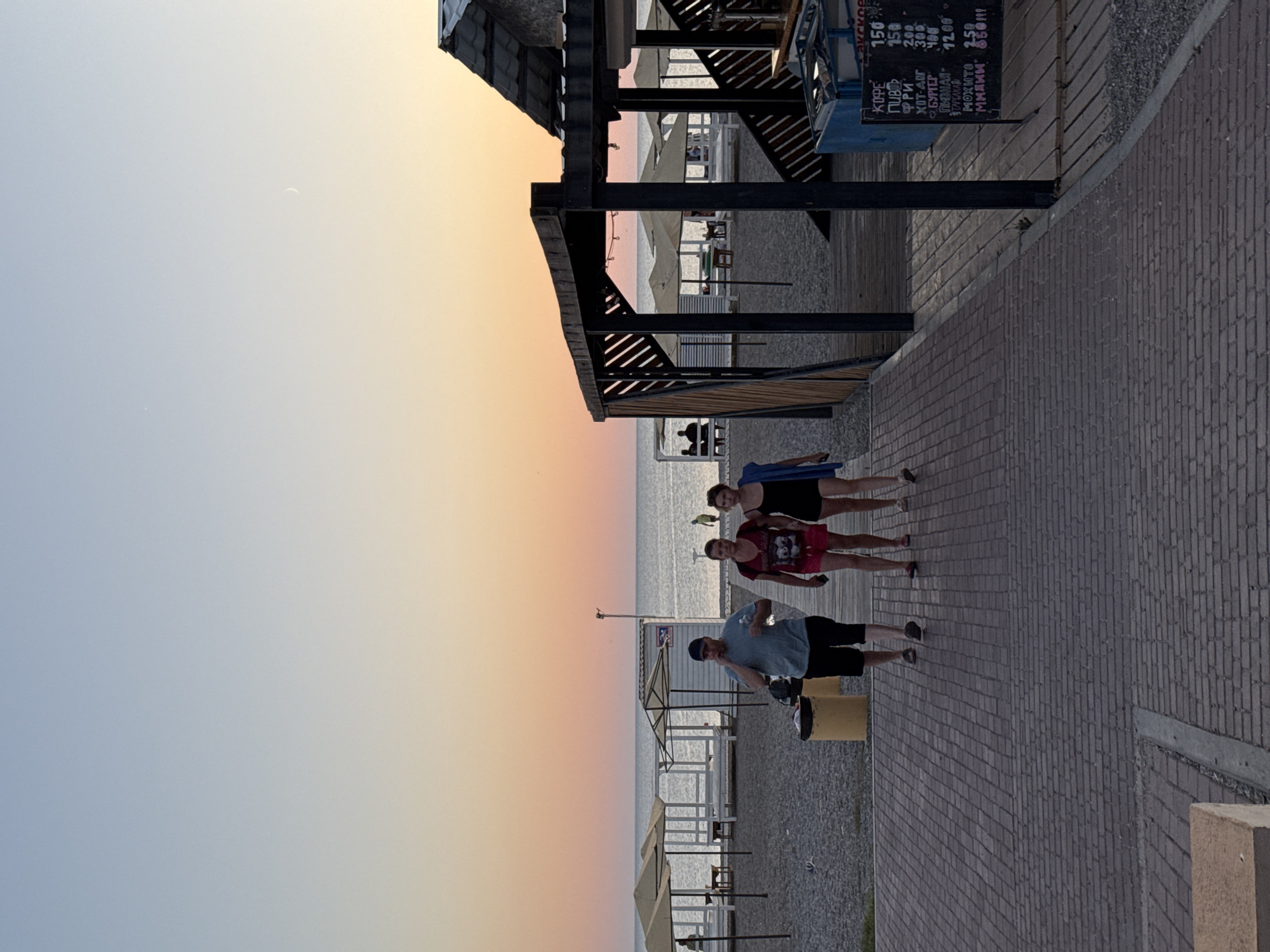 A group of three people walk along a paved pathway leading towards a beach. The person on the left is a man wearing a blue t-shirt and black shorts, holding a phone to his ear. In the middle is a woman in a red t-shirt and pink shorts. To her right is another woman in a black swimsuit, carrying a blue towel. The scene is set at dusk with a clear sky transitioning from pale blue to orange and pink hues. In the background, there are several beach umbrellas and white structures. To the right of the pathway, a wooden structure with a slanted roof is visible, and a chalkboard menu is propped against it, displaying prices in Russian. The ground is a mix of paved bricks and gravel, indicative of a coastal boardwalk or promenade. The overall setting appears to be a public beach area in Novofedorovka, Ukraine.