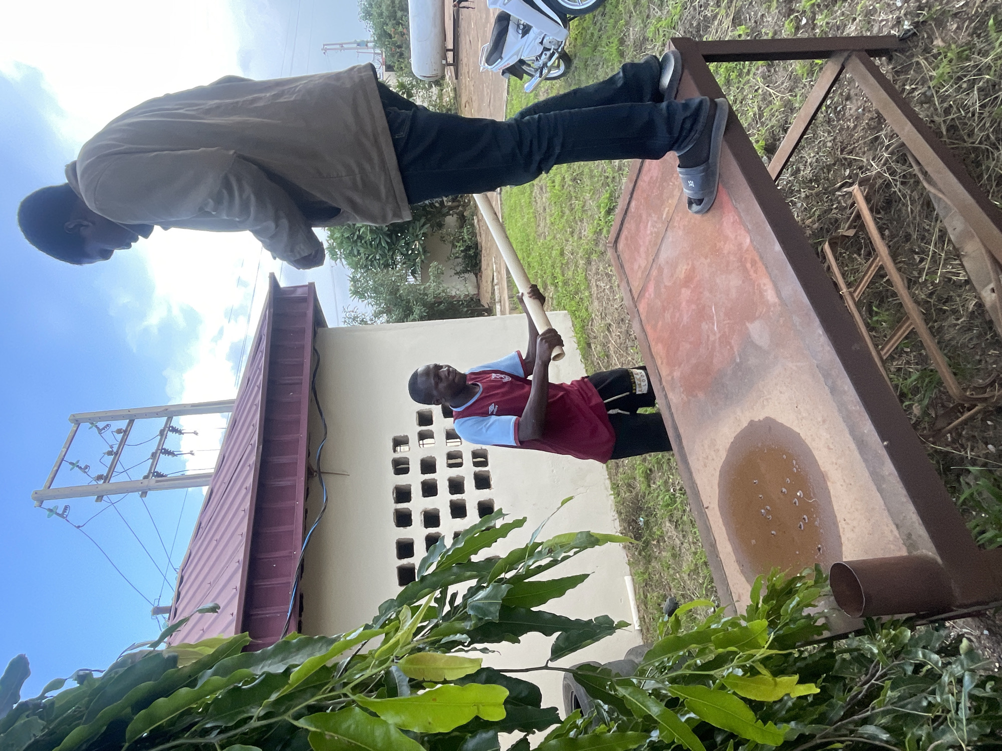 In the city of Jos, Nigeria, two young men are engaged in an activity on an outdoor metal table. One man, wearing a maroon and light blue jersey and black shorts, stands on the ground to the left, holding a light-colored cylindrical object. The second man, wearing a tan jacket, dark blue jeans, and grey sandals, stands on top of the metal table to the right, facing away from the camera. The table has a reddish-brown, worn surface, with a puddle of brown liquid and small white specks on it. A brown cylindrical object is attached to the side of the table.
The environment is grassy with some dry patches. Behind the men, a cream-colored building with a maroon roof and a wall featuring a pattern of square openings is visible. Electrical poles with wires are seen against a cloudy sky. To the right of the building, a white motorcycle and a large industrial pipe are present. Lush green foliage hangs from the left side of the frame, partially obscuring the scene.
