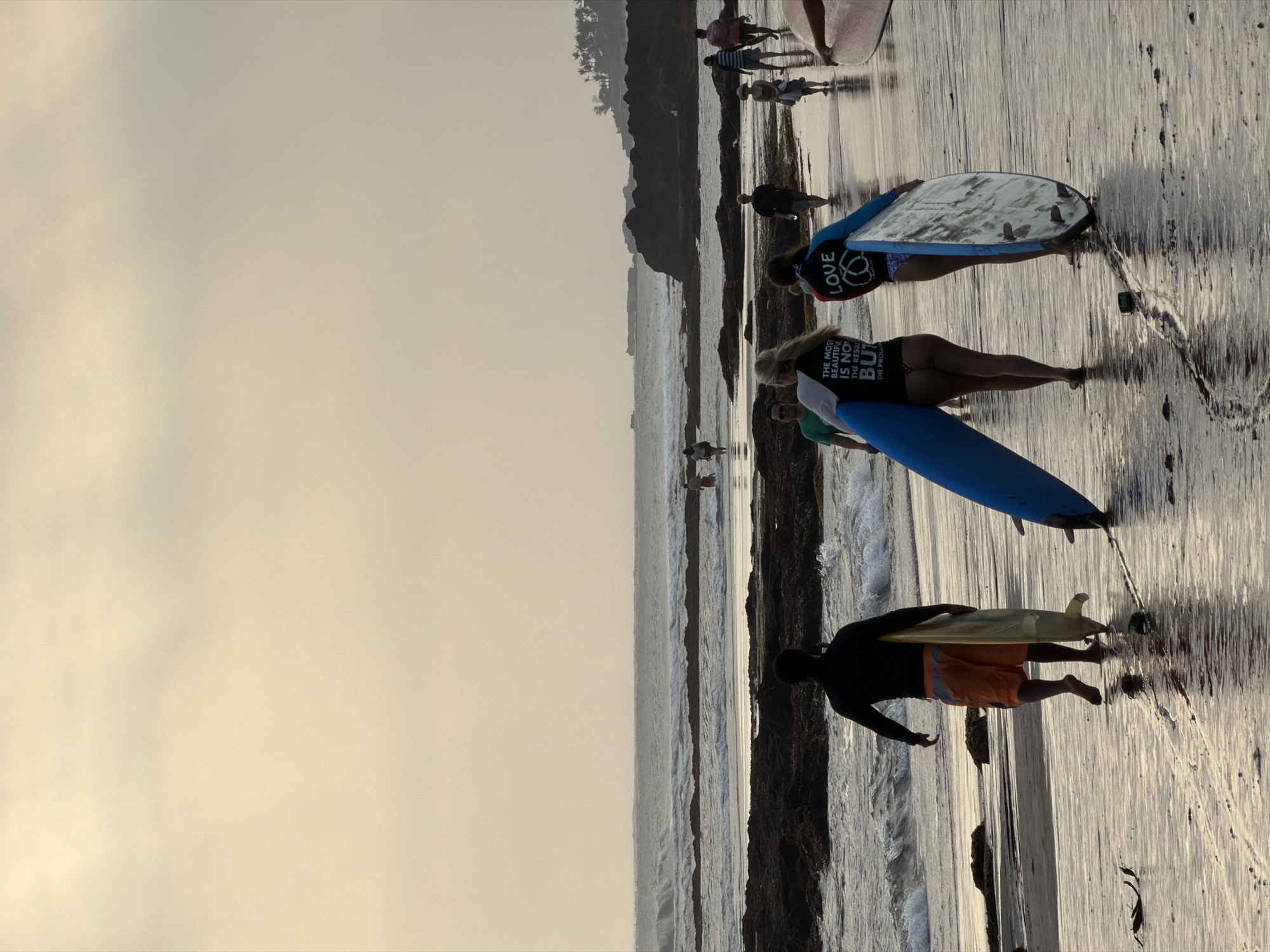 At dusk on the beach in North Kuta, Bali, Indonesia, a group of people walk along the wet sand. The tide is low, revealing rocks and pools of water. A surfer, silhouetted in a black long-sleeved shirt and orange board shorts, carries a yellow surfboard. Further ahead, two women walk with surfboards. The woman on the left wears a black shirt with white text that reads, "THE MOST BEAUTIFUL IS NOT THE RESUL BUT THE PROCESS." The woman on the right wears a blue and red long-sleeved shirt with "LOVE" printed on it. Other beachgoers are visible in the background, walking towards and away from the ocean. The sky is overcast with a pale yellow hue.