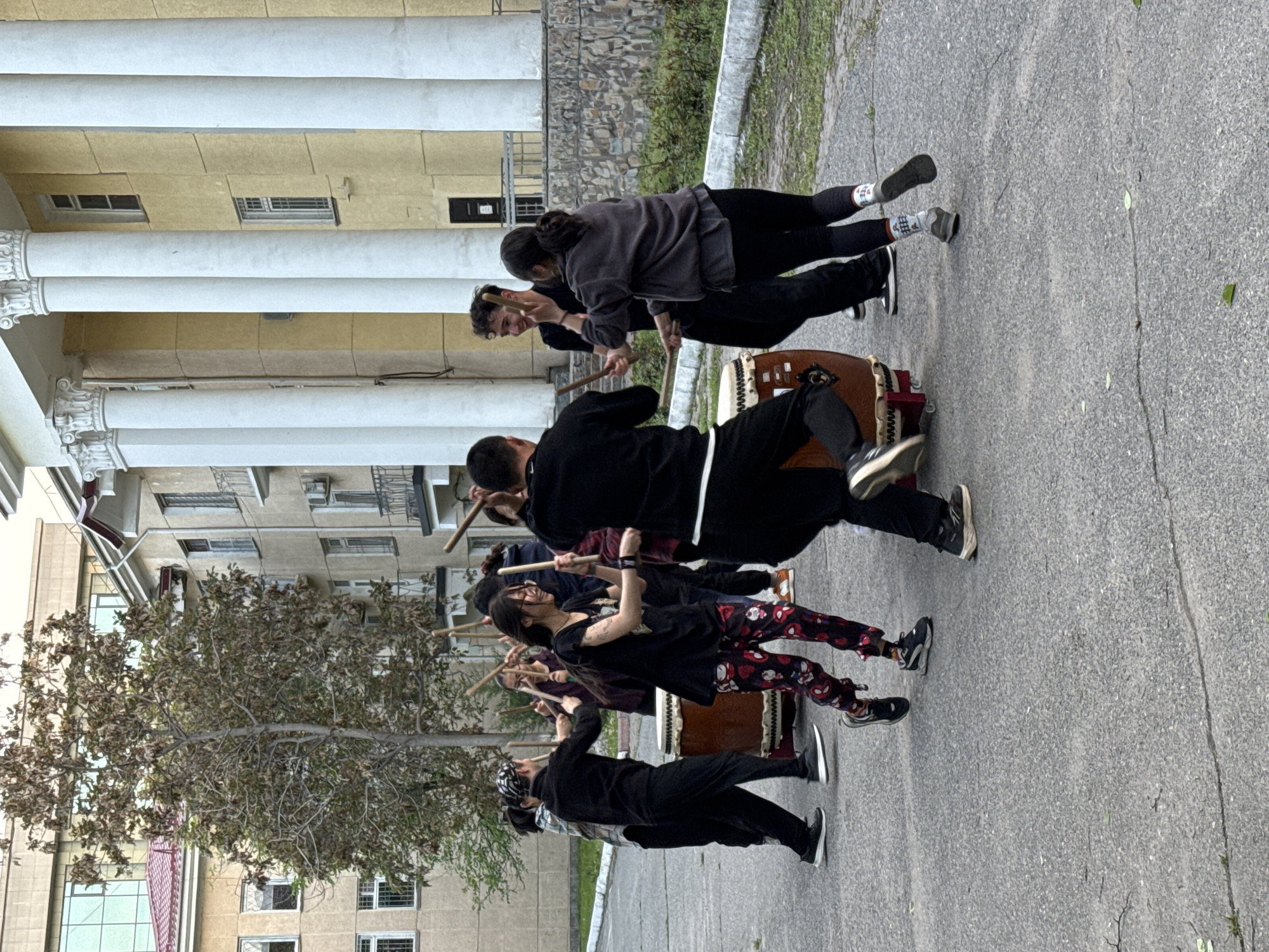 A group of people are playing taiko drums on an asphalt paved surface, in what appears to be a public area in Bishkek City, Kyrgyzstan. The group is arranged in a row, with each person holding drumsticks poised to strike a large, brown taiko drum. The people are dressed in casual clothing, with some wearing dark colors and others wearing patterned pants. Buildings with white columns are visible in the background, along with trees, and other architectural structures. The image orientation is vertical.