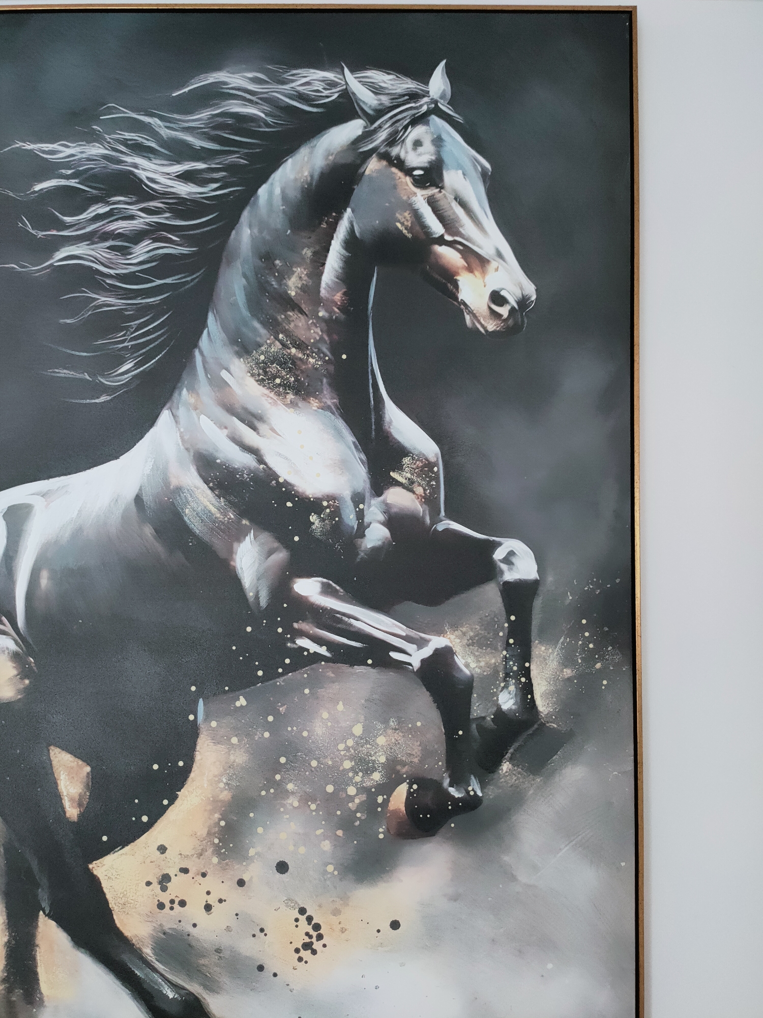 This is a medium shot, eye-level photograph of a framed painting of a black horse. The painting is dominated by the horse, which is depicted in mid-gallop, rearing up on its hind legs. Its mane is flowing dynamically in the wind, and the horse's body is highlighted with streaks of light and gold flecks that suggest movement and power. The background is dark and moody, with smoky textures. The painting is framed in a thin, golden frame. To the right of the painting, a white wall is visible. The photograph appears to be taken indoors. The painting's subject matter and style suggest it might be a decorative piece in a residential or commercial space. The image does not contain any information about Mrezga, Tunisia.