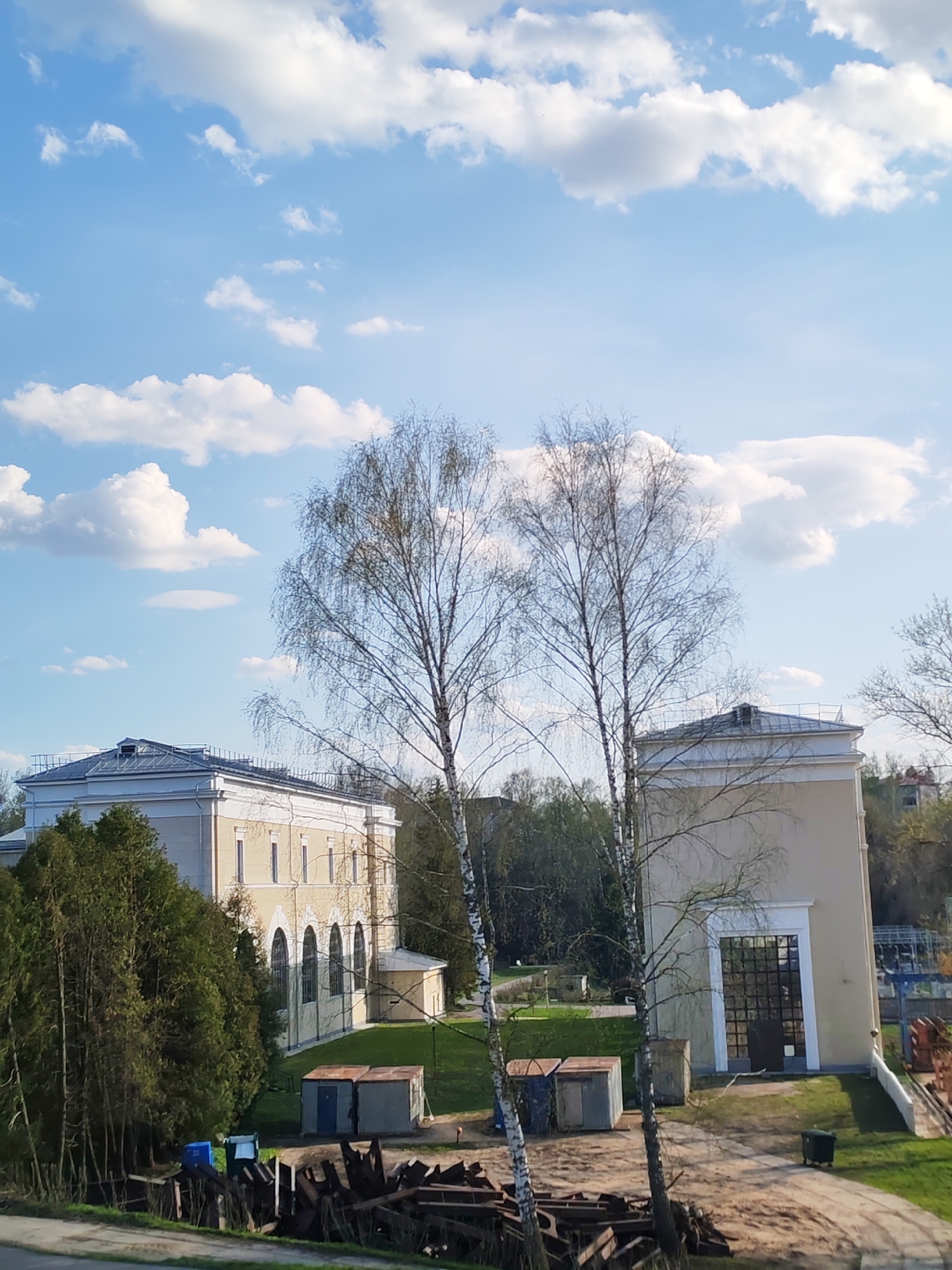 The image depicts a daytime scene in what appears to be a suburban or semi-rural area of Водник Снт, Russia. The sky is blue with scattered white clouds. Two buildings stand prominently in the midground. The building to the left is a two-story structure with arched windows and a light-colored facade. To the right is a taller, narrower building with a large glass door. Between the two buildings is a grassy area with several smaller structures or sheds. Two bare trees are positioned in the foreground, partially obscuring the view of the buildings. In the very foreground, there's what seems to be a pile of debris or construction materials. The overall impression is of a quiet, somewhat industrial or utility-focused section of the city.