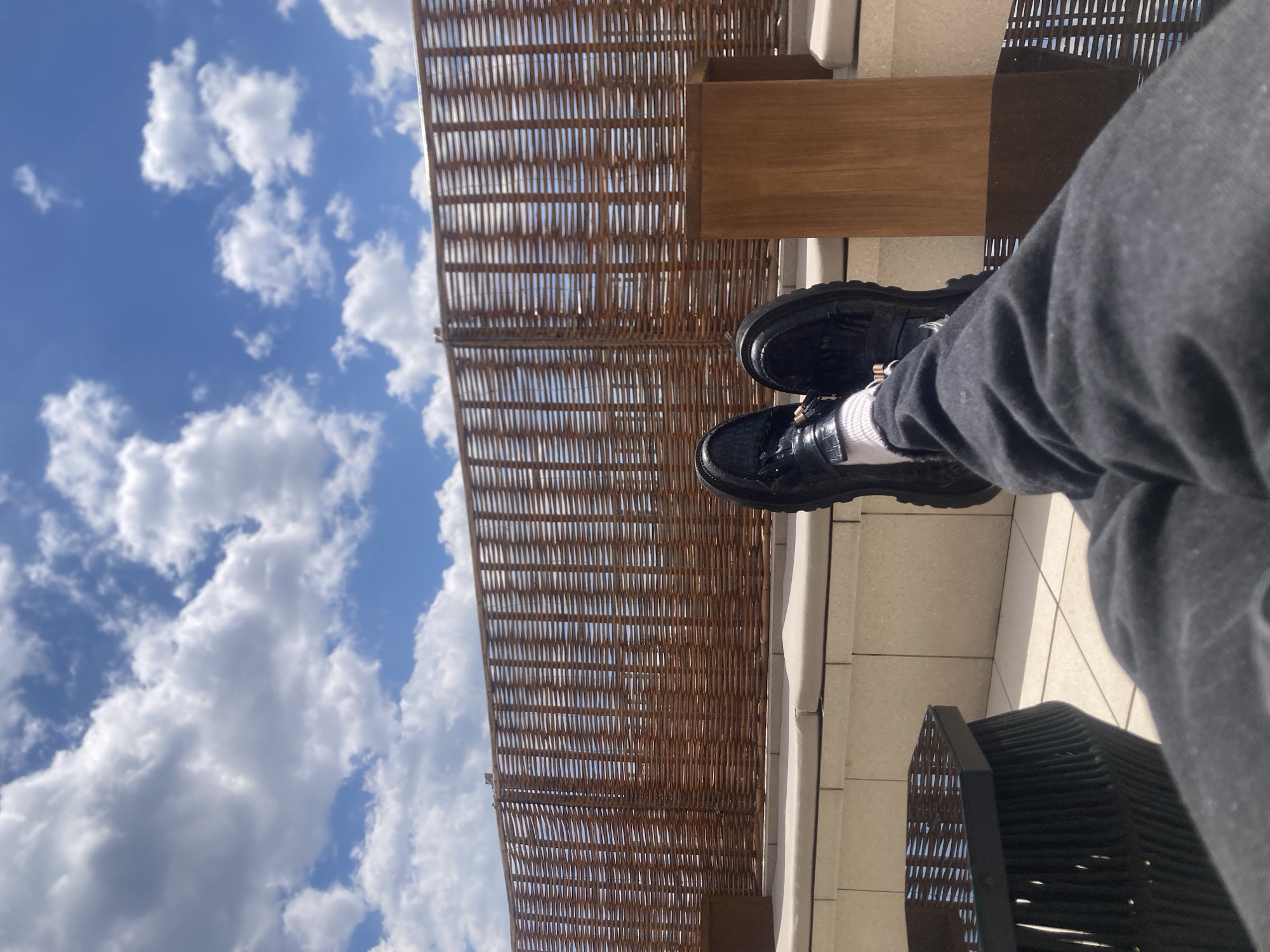 A low-angle, outdoor shot shows a person's legs and feet in black shoes and white socks, with the sky and clouds visible in the background. A woven structure is positioned above, possibly a pergola or canopy. The environment suggests a rooftop or terrace setting, featuring tile flooring and a glimpse of outdoor furniture. The picture does not provide any specific elements that would permit the identification of the location within the city of Barcelona, Spain.