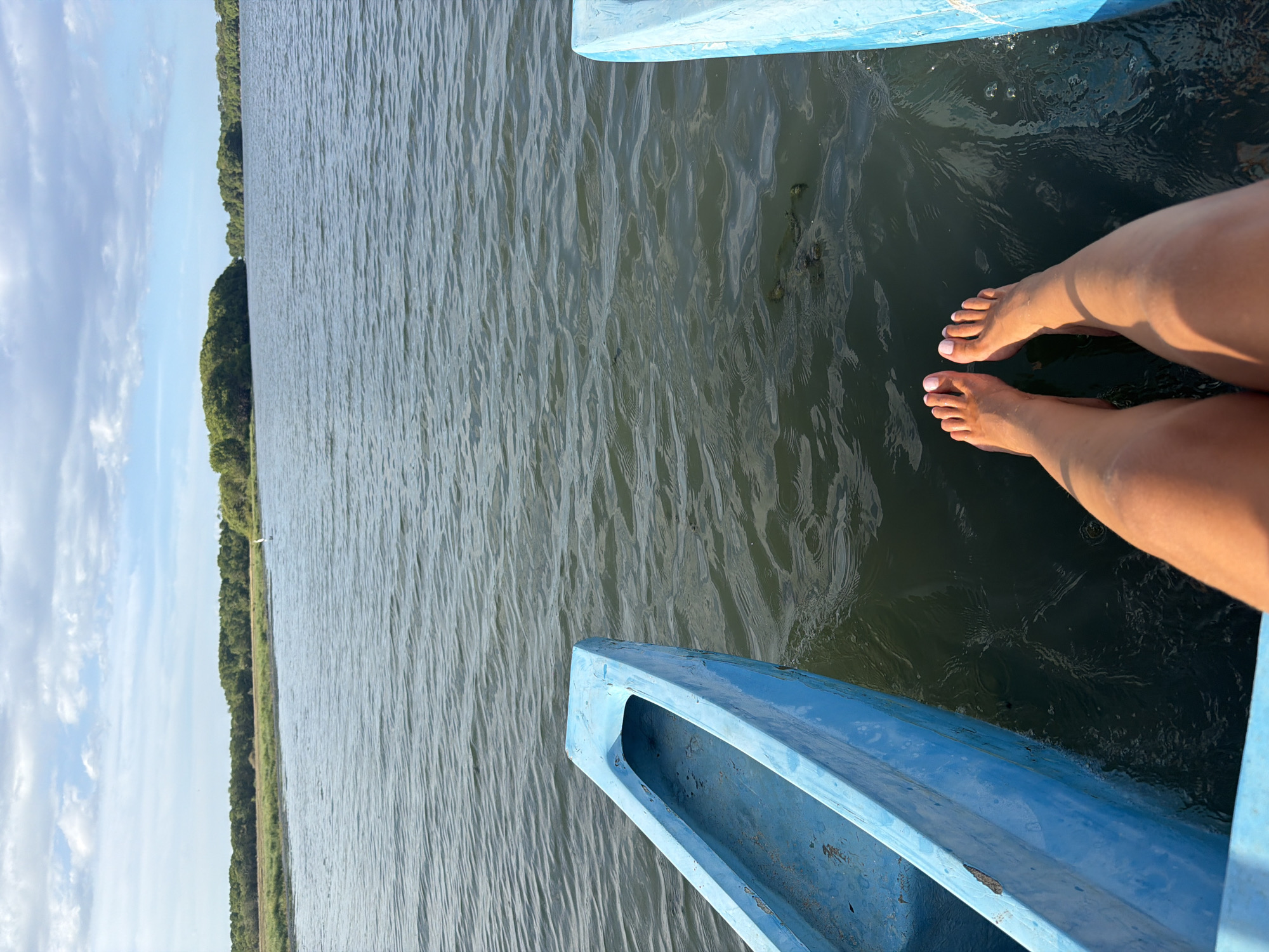 A pair of bare feet with pink nail polish rests on the edge of a bright blue boat, with the toes extending over the water. The boat, which appears to be a canoe or similar vessel, is positioned in a body of calm, dark water with ripples on the surface. In the background, a lush green shoreline with trees and vegetation is visible under a partly cloudy sky. The location is identified as Pottuvil, Sri Lanka.