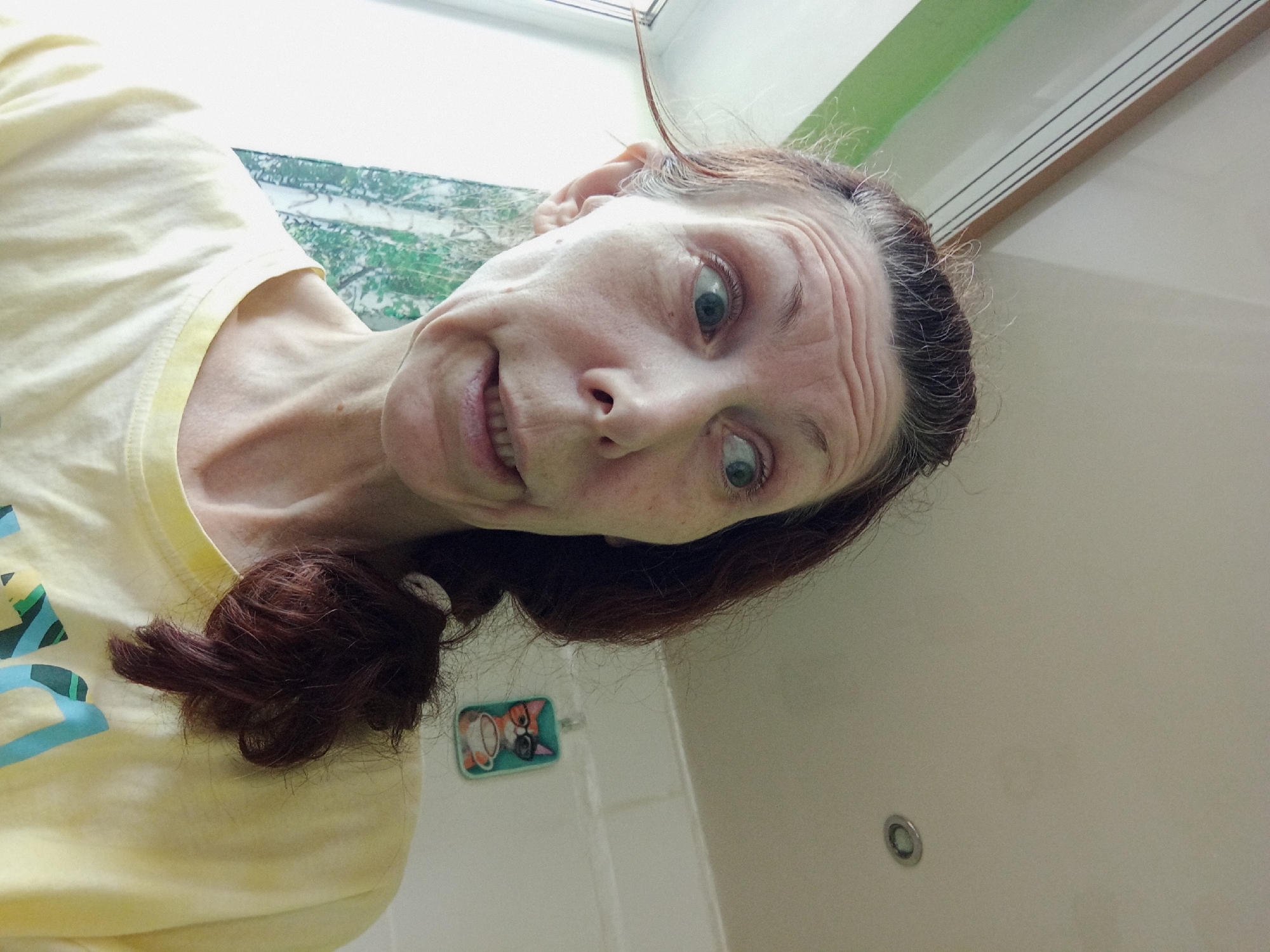 A close-up, over-the-shoulder selfie taken indoors shows a woman with light skin, auburn hair in a low ponytail, and blue eyes. She is wearing a yellow t-shirt with a graphic design and appears to be looking off to the side with a surprised expression. The background includes a window with a green curtain, a ceiling light, and a small decorative plaque featuring a cat. There are no elements in the image that would allow for the determination of the location within the city of Blagoveshchensk, Russia.