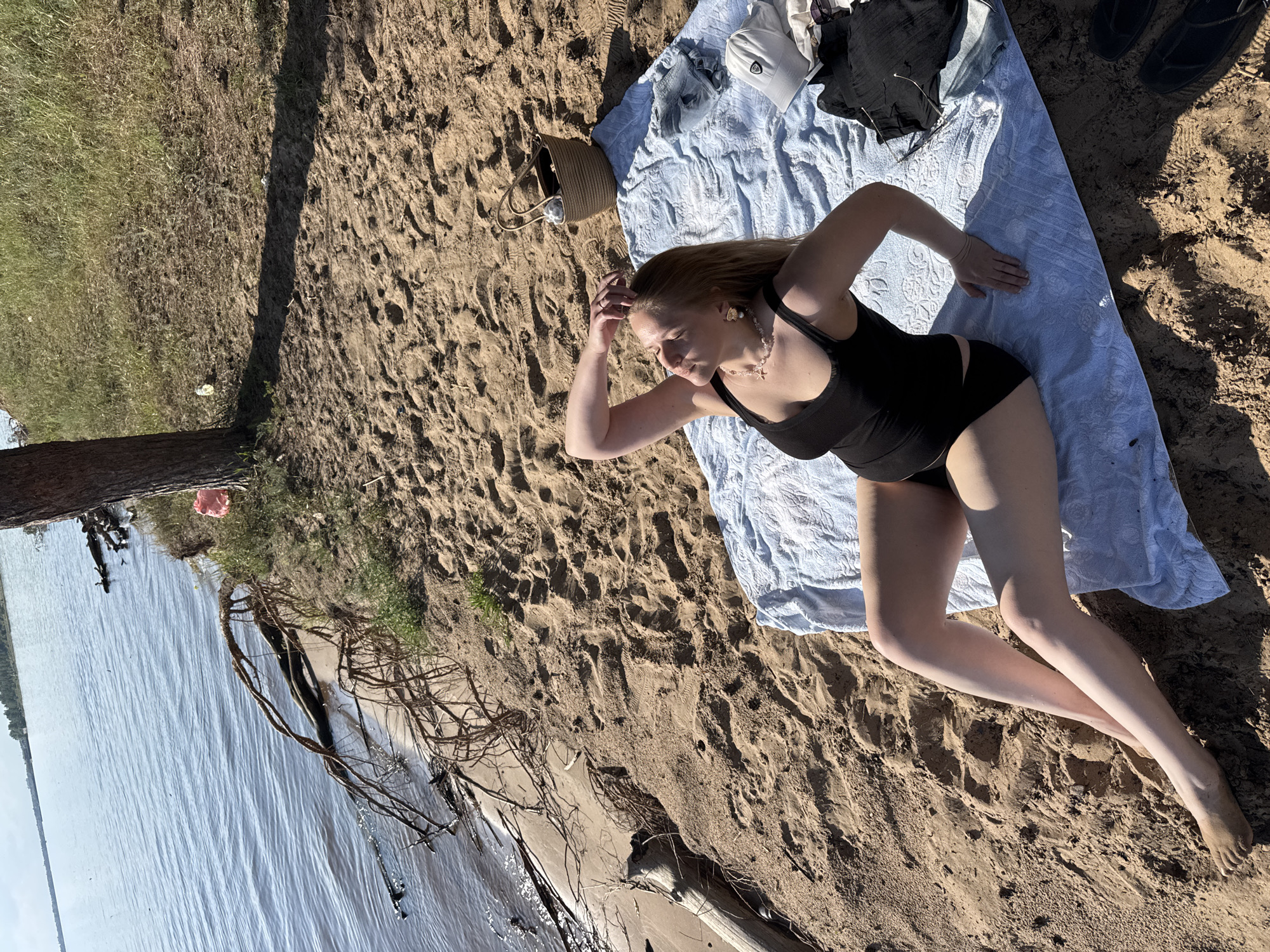A woman in a black swimsuit lies on a blue towel on a sandy beach. Her eyes are closed, and she is resting her head on her hand, suggesting relaxation. Several items are scattered around her on the towel, including a straw bag, a white baseball cap, and some dark clothing. The scene is illuminated by sunlight, casting shadows on the sand, indicating it is daytime. The presence of the water and sand suggests an outdoor, possibly recreational setting. The woman's posture and the tranquil atmosphere hint at a leisurely activity like sunbathing.
