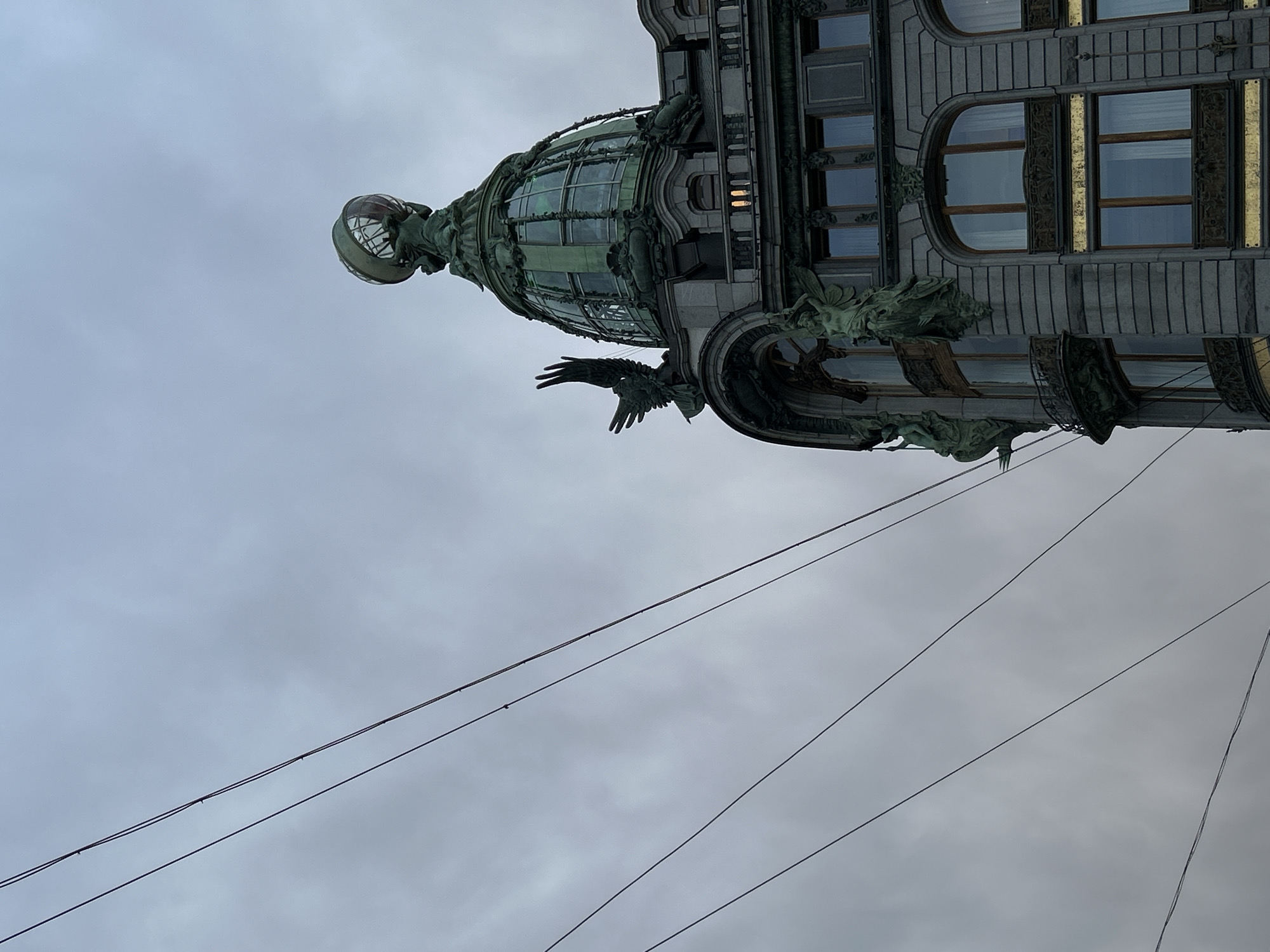 A low-angle shot showcases the architectural details of the Singer House building in St. Petersburg, Russia, under a cloudy sky. The building features ornate decorations, including a glass dome topped with a globe, and sculpted eagles. Power lines extend across the foreground. The building's facade includes multiple windows and decorative elements. The overall atmosphere is overcast.