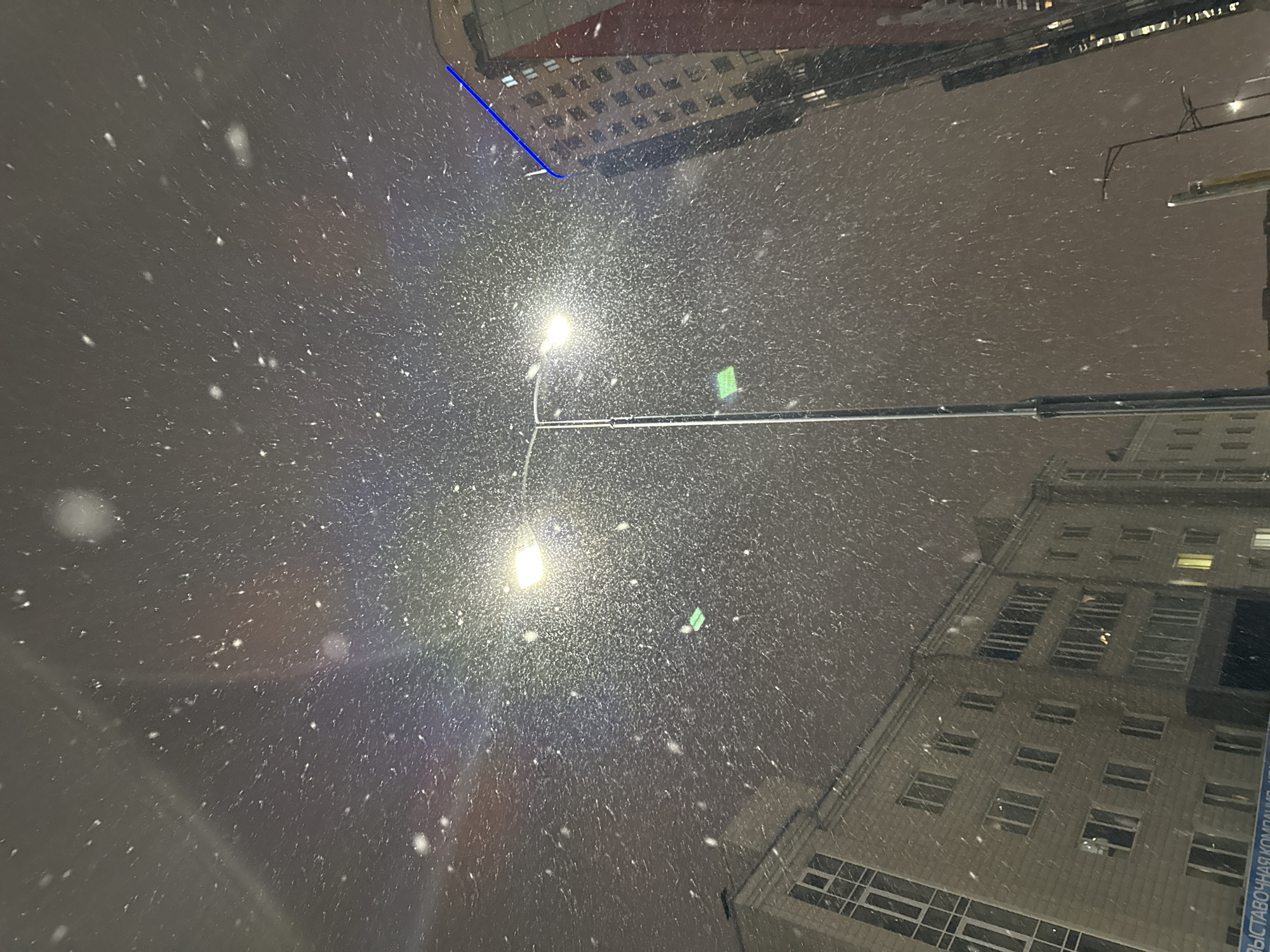 The image, shot from a low angle, depicts a heavy snowfall at night in an urban area. Two tall buildings frame the scene on the upper left and lower right sides, their illuminated windows visible through the falling snow. A street lamp shines brightly in the center of the image, its light diffused by the snow. A horizontal beam stretches across the middle of the frame, possibly a support structure for the lamp or other street fixture. The sky is a dark, overcast gray. Based on the presence of Cyrillic script on the building to the lower right, this scene is likely located in Krasnoyarsk, Russia. There are no people visible in the photograph.
