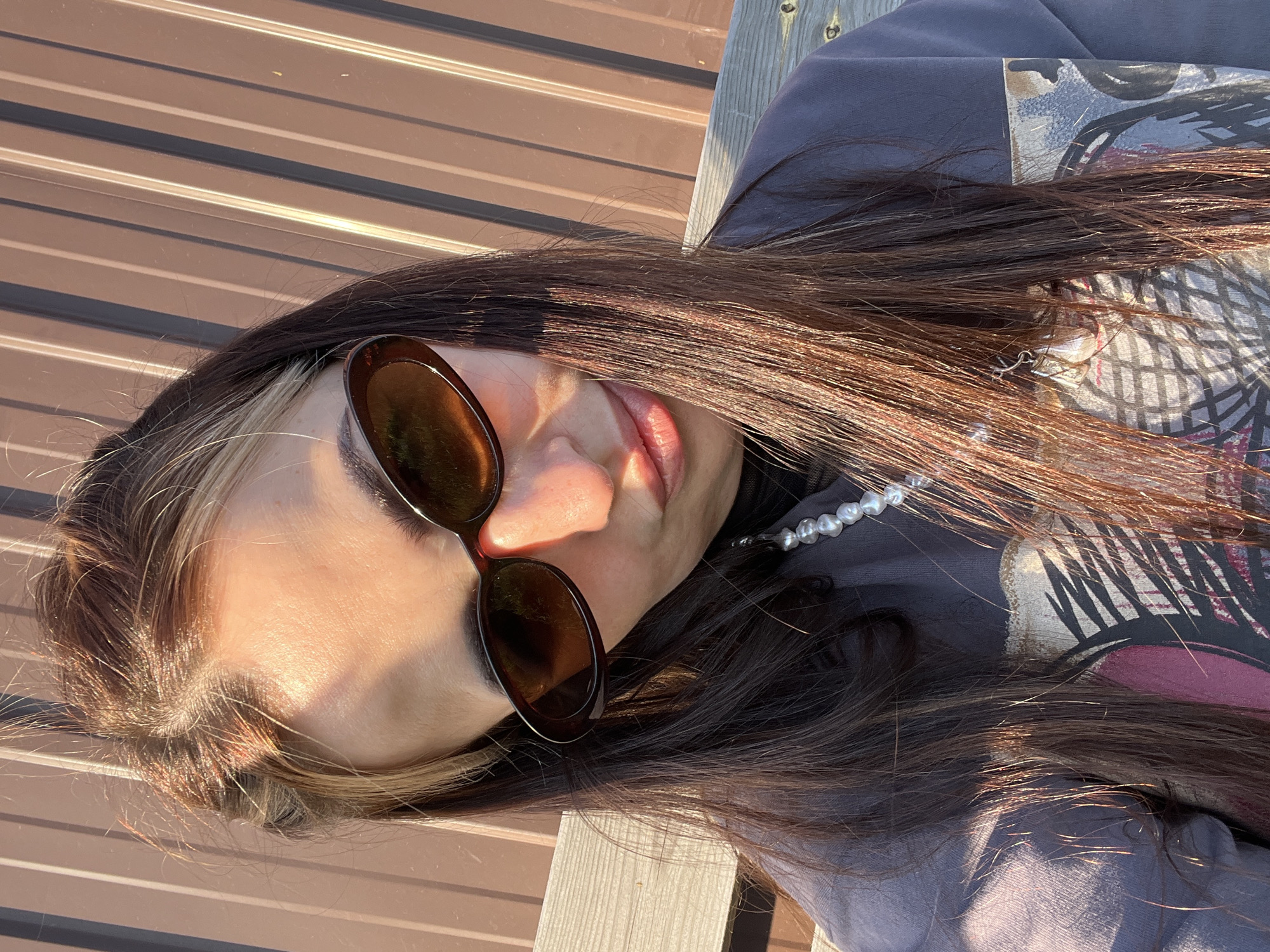 A portrait of a woman is oriented at an oblique angle, set against a backdrop of horizontally aligned brown panels. The woman, positioned in the foreground, wears tinted sunglasses and a string of pearls, with her long brown hair cascading around her face and shoulders. She is dressed in a grey shirt with a graphic print. The scene appears to be bathed in natural sunlight, creating strong shadows on her face and the background. The setting and location are unidentified.
