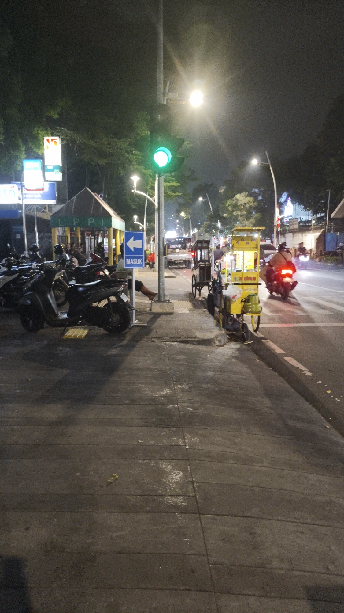 The image depicts a street scene at night in South Jakarta, Indonesia, likely near the "Gedung Nyi Ageng Serang" building, as suggested by the visible signage. A green traffic light is illuminated, indicating that vehicles are permitted to proceed. Several parked motorcycles are visible on the left side of the frame, and a person is partially visible sitting near them, with their legs extended.
To the right of the motorcycles, a blue directional sign with a white arrow pointing left and the word "MASUK" (Indonesian for "Entrance") is mounted on a pole. Further to the right, a yellow food cart, possibly selling snacks or drinks, is positioned on the sidewalk. Another cart, darker in color and with spoked wheels, is visible behind it.
On the street, a motorcycle with its taillight illuminated is moving away from the viewer. In the background, a truck with its headlights on is also on the road. The environment suggests a busy urban setting with streetlights and trees visible in the dark background. The pavement is made of rectangular concrete slabs, some of which appear worn and marked. The overall lighting is dim, characteristic of nighttime illumination.