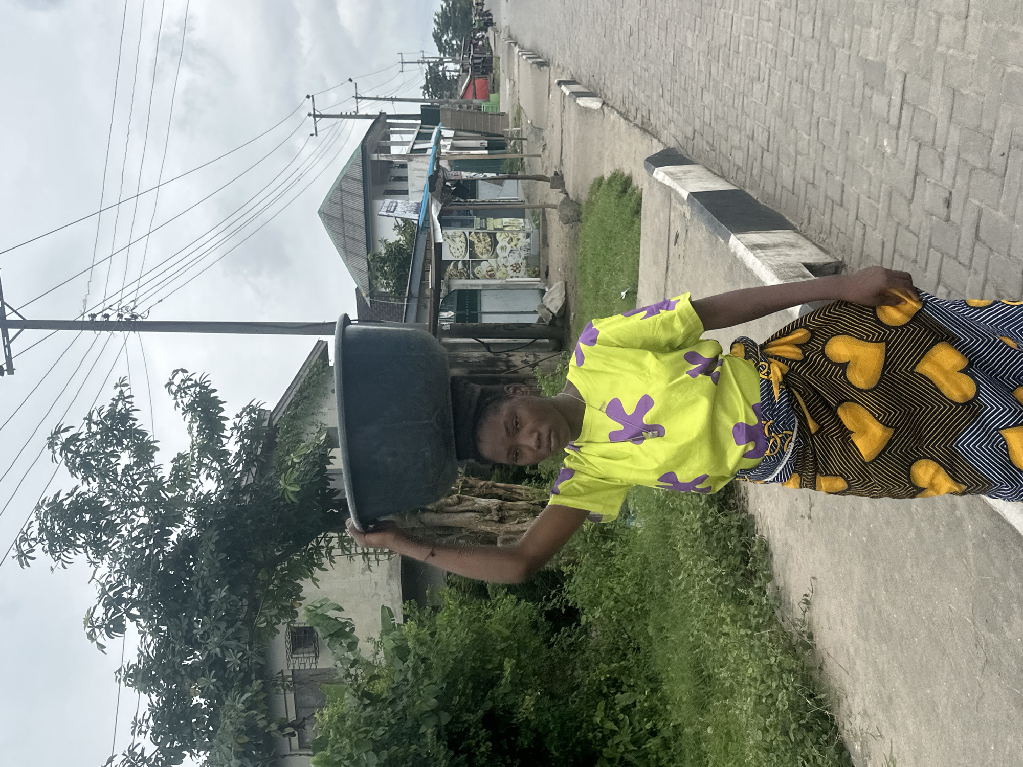 In Badagry, Nigeria, a young woman walks along a cobblestone street carrying a large black basin on her head. She wears a vibrant lime green t-shirt with purple floral patterns and a colorful patterned skirt. The street is lined with buildings, including what appears to be a small restaurant or shop with food displays, and various trees and greenery. Utility poles with numerous wires stretch across the cloudy sky, indicating an urban or semi-urban setting. The scene captures a moment of daily life, with the woman likely transporting goods or water.