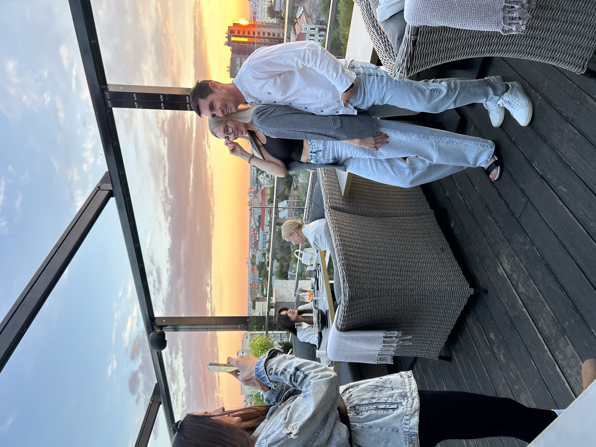 This image depicts a group of people on a rooftop patio during sunset. The sky is filled with warm hues of orange and yellow, indicating either sunrise or sunset. In the foreground, two individuals, a man and a woman, are posing for a photo. The man, wearing a white long-sleeved shirt and light-wash jeans, smiles at the camera. The woman, in a grey cardigan over a black top and light-wash jeans, is also looking towards the camera. They are standing on a dark wooden deck, with outdoor furniture and a railing visible.
Behind them, other people are seated at tables, enjoying the view. One person is looking at their phone. The background shows a cityscape with buildings and trees, suggesting an urban environment. The overall atmosphere appears relaxed and social, possibly a gathering at a restaurant or bar. The time of day is clearly sunset, given the sky's colors. There is no visible text in the image.