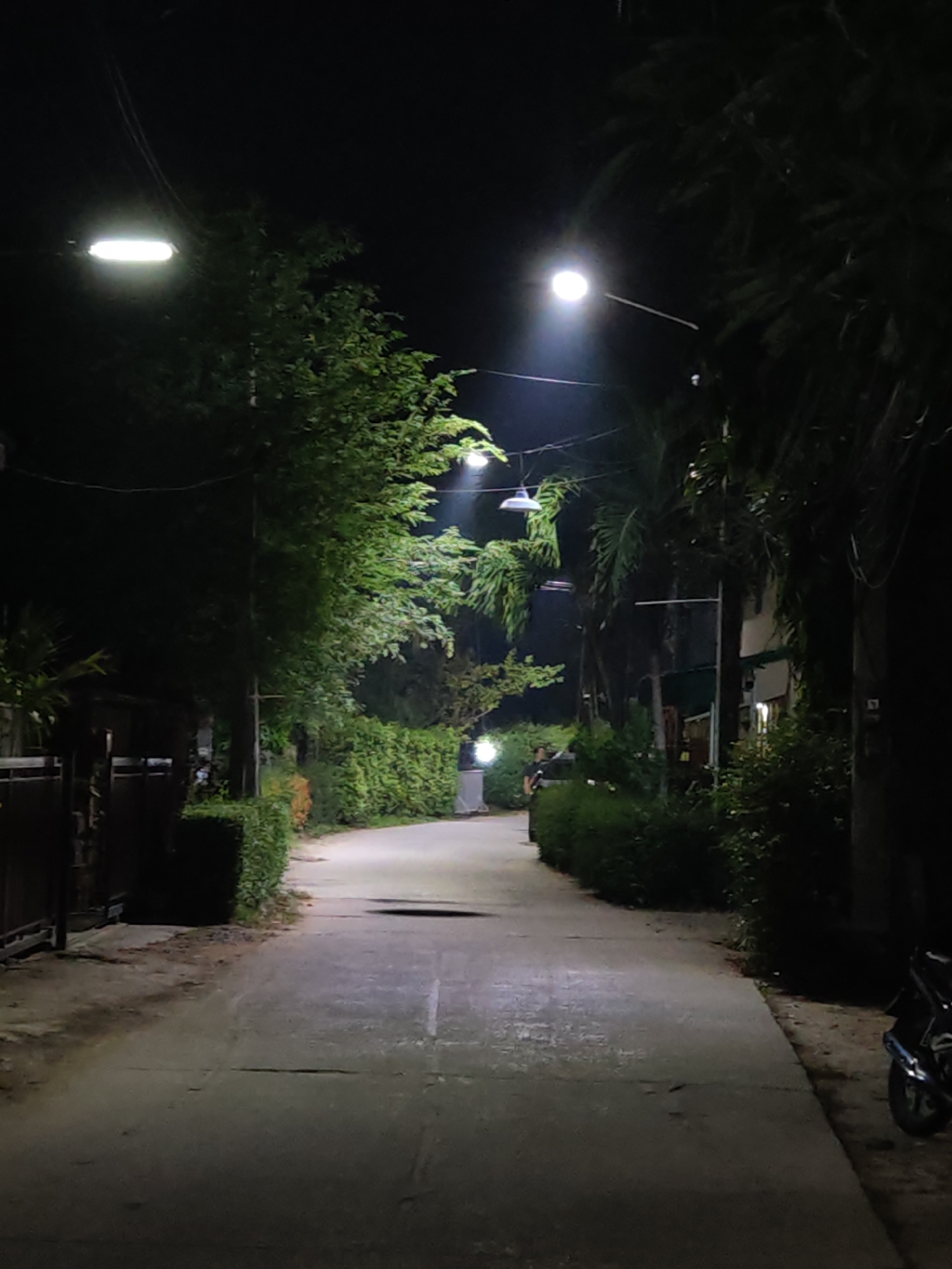 The image shows a dark, narrow street in Baan Bo Phut, Thailand, illuminated by streetlights. Trees and dense foliage line both sides of the road, casting shadows. A paved path curves into the distance, bordered by manicured hedges and a fence on the left. A motorcycle is parked on the right side of the road. The scene appears deserted, with no people visible.