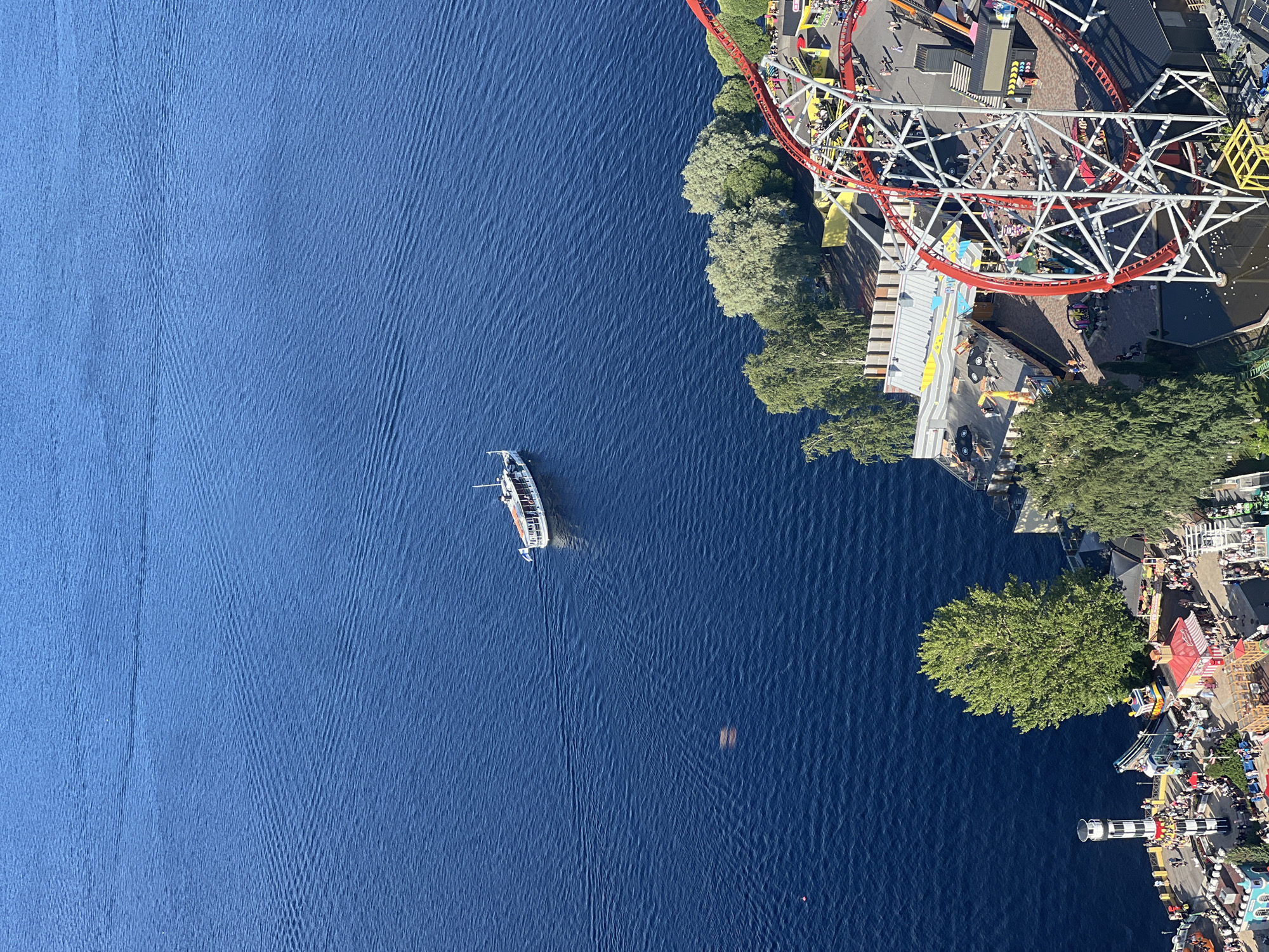 This aerial image captures a view of Tampere, Finland, focusing on a section of the city bordering a large body of water. In the foreground, a prominent red roller coaster with a white support structure is visible, winding through an amusement park area. Several buildings with colorful roofs and awnings are scattered throughout the park, alongside trees and pathways where numerous people are seen walking and congregating. A smaller body of water, possibly a pond or a decorative feature, is present near the base of the roller coaster.
Further out, a white passenger boat with an orange-striped awning is sailing on a vast expanse of deep blue water, leaving a faint wake. The water stretches to the horizon, where it meets a clear blue sky. The shoreline area features a lighthouse-like structure with black and white stripes, and more trees line the edge of the water. The overall impression is a vibrant summer day at an amusement park situated by the water.