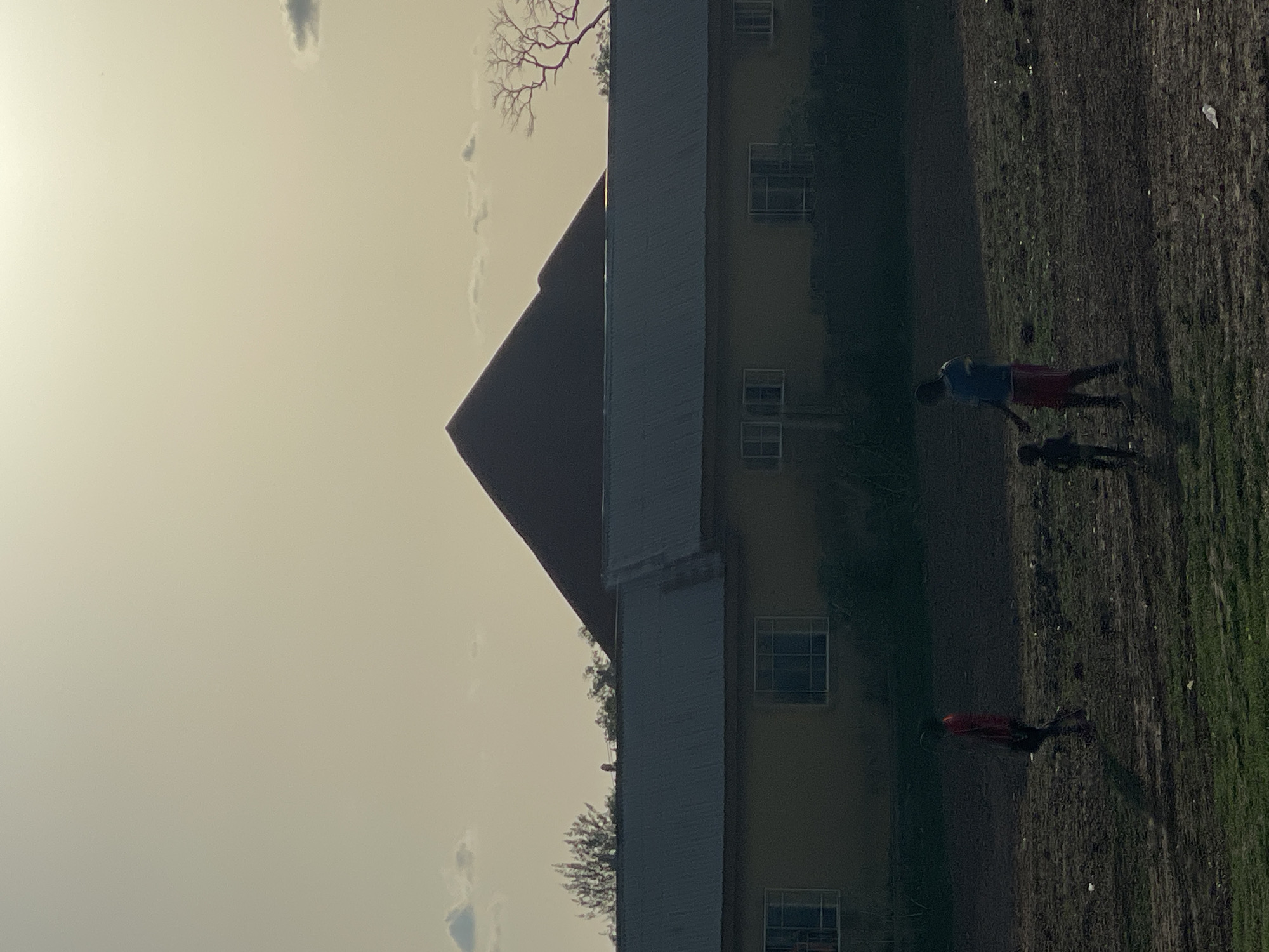 A building with a red roof and beige siding stands next to a grassy field. Two people are walking in the field. Another person is also walking near the building. The sky is bright. The location is Lokoja, Nigeria.