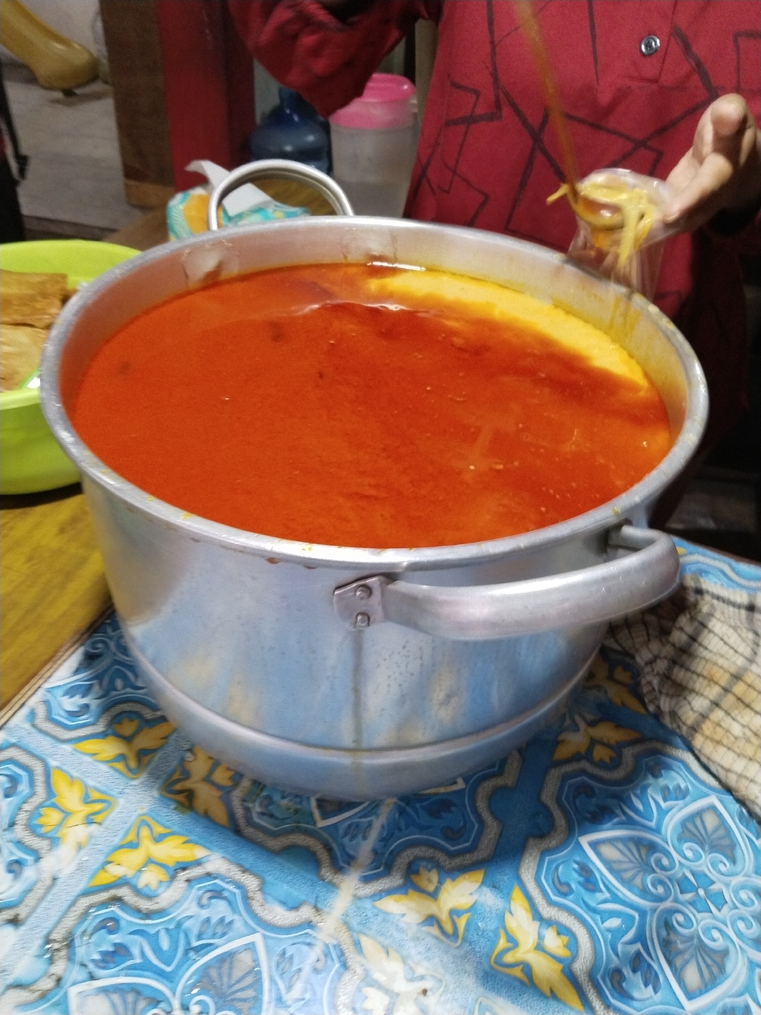 A close-up, overhead view captures a large, aluminum pot filled with a vibrant, orange-red soup. The soup has a slightly oily sheen and appears to have a smooth texture with a lighter yellow layer on top. Two silver handles are attached to the sides of the pot. The pot sits on a blue and yellow patterned tablecloth. To the left of the pot, a bright green bowl contains what appear to be fried tofu. In the background, a person wearing a red patterned shirt is visible, holding a plastic bag filled with yellow noodles, seemingly in the process of serving them. A pink bottle and a blue bottle are also visible in the background. The scene suggests a food preparation or serving area, possibly a street food stall or a home kitchen.