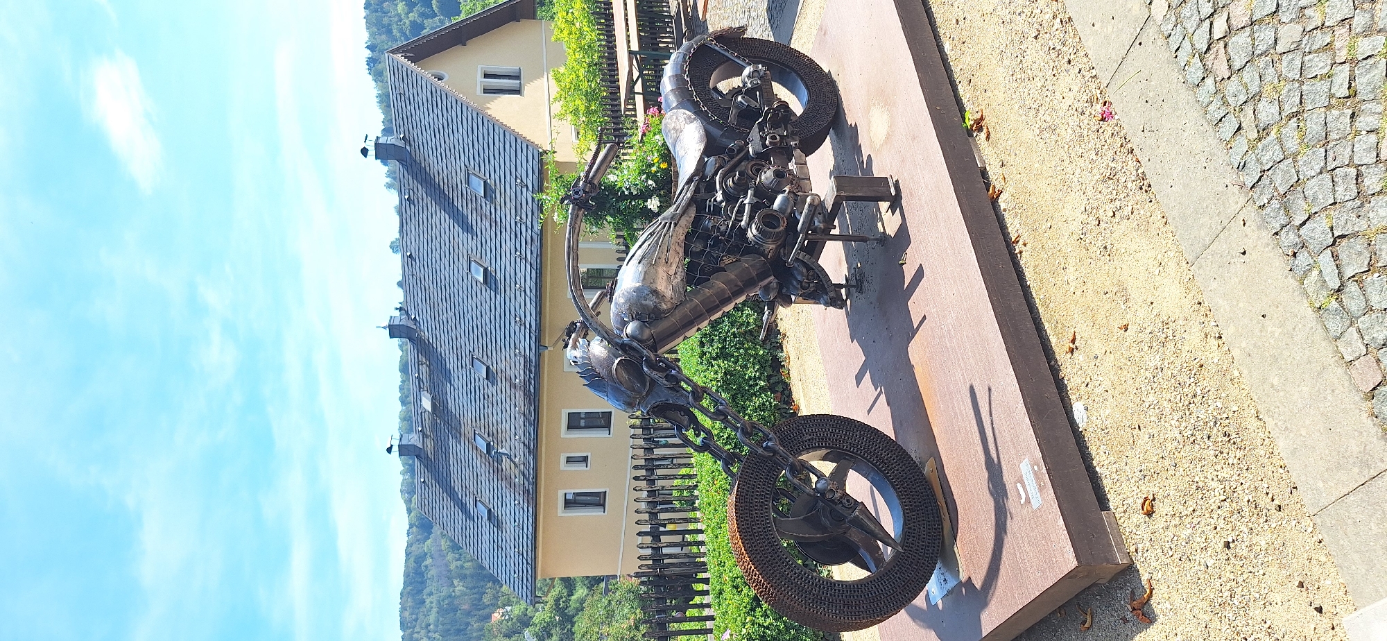 This is a full-shot, eye-level, vertical-format photograph of a metal sculpture of a motorcycle displayed outdoors. The sculpture is metallic and appears to be made from recycled materials, with visible chains and various metal components forming the engine, frame, and wheels. The front wheel is on the left and the rear wheel is on the right. The motorcycle sculpture is placed on a large, dark brown, rectangular platform, which is elevated slightly from the ground. The ground around the platform is covered with gravel and fallen leaves.
Behind the sculpture, there is a dark wooden fence, and behind the fence is lush green foliage, including bushes and trees. Further in the background, there is a light yellow house with a dark grey, sloped roof featuring several dormer windows. The house is situated on a gentle slope, with more trees and greenery visible on the hillside behind it. The sky is bright blue with scattered white clouds.
The location is identified as Zschopau, Germany. Specifically, the motorcycle sculpture is located in front of the Hotel am Schloss in Zschopau. The Hotel am Schloss, also known as the Jagdschloss (Hunting Lodge), is a historical building in the town.