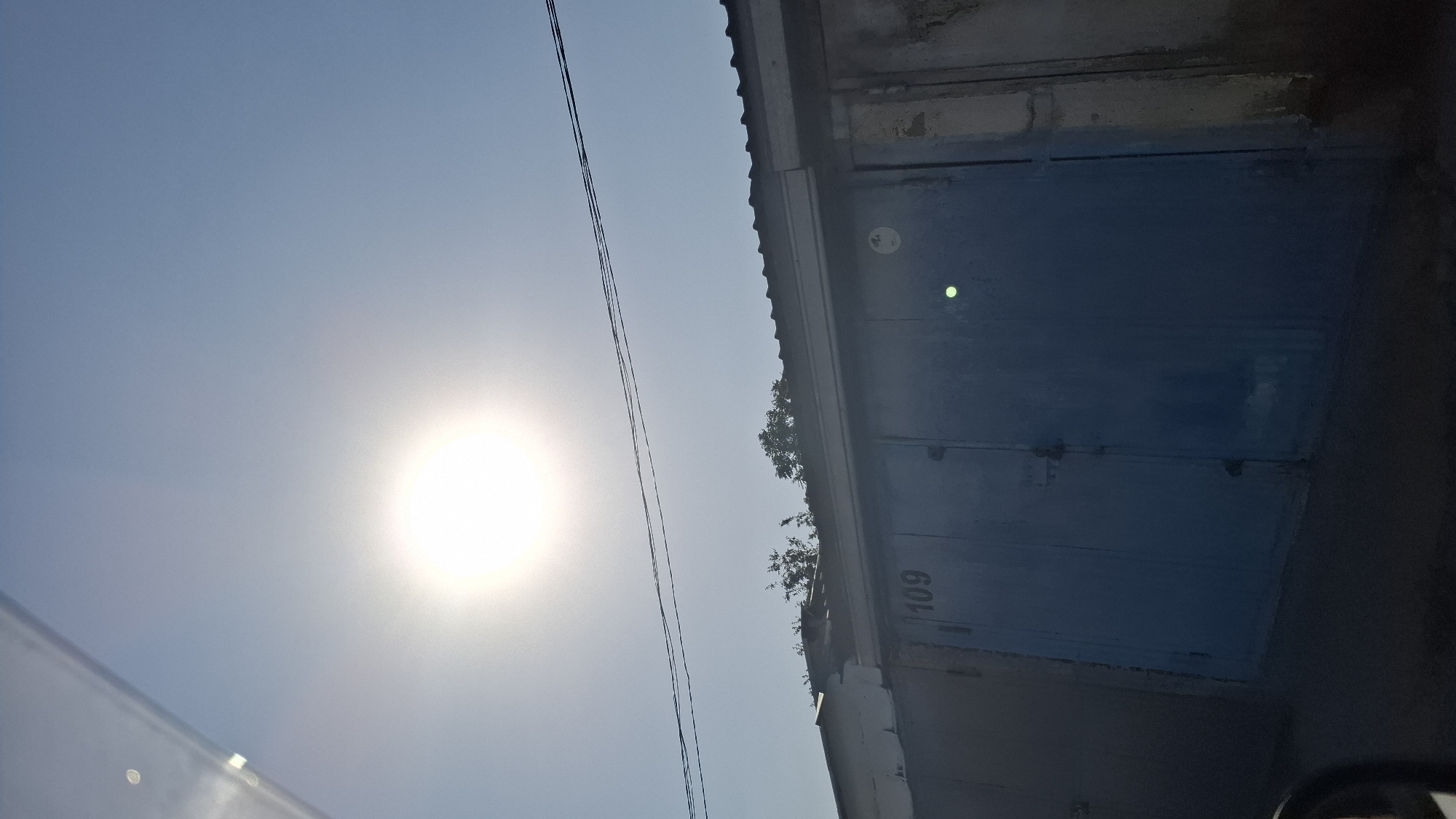 The image shows a clear blue sky with a bright sun in the upper left quadrant. The sun's rays are diffused, creating a hazy effect. Below the sky, a row of garage doors is visible. The doors are a faded blue color, and some appear to be made of metal. A number "109" is visible on one of the doors. The garages have a dark, corrugated metal roof. Trees are visible behind the garages. The image appears to have been taken on a sunny day in Shymkent, Kazakhstan, likely in a residential or industrial area with multiple garages. The angle of the sun suggests it is midday.