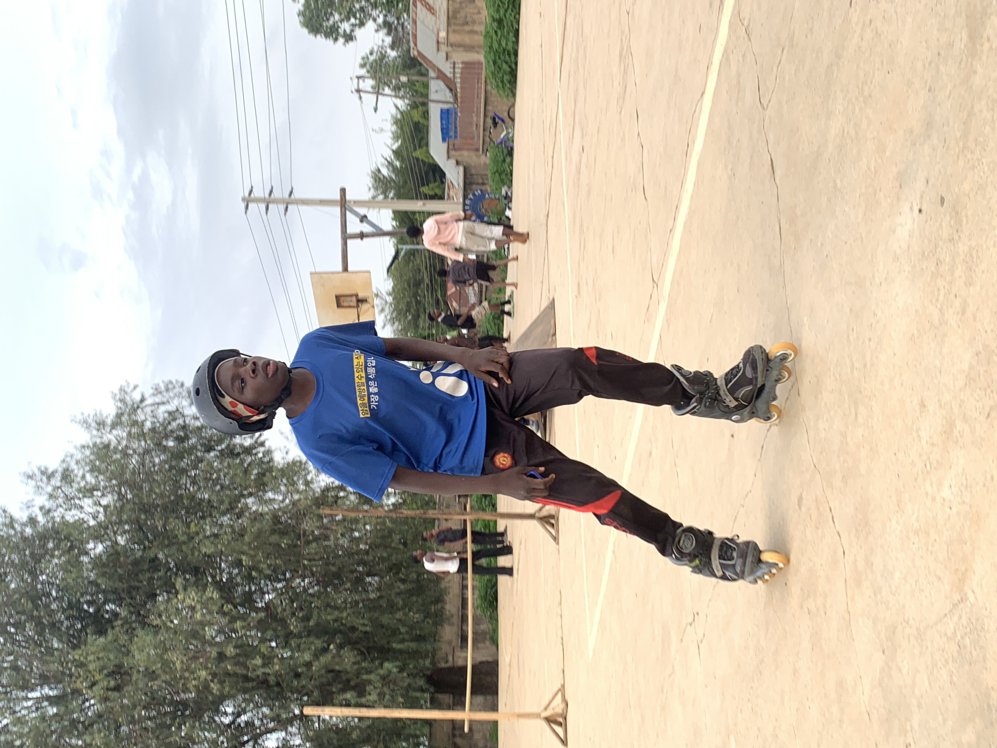 A young Black male, wearing a helmet, blue T-shirt with Korean script, and black pants with red accents, is standing on rollerblades on a concrete surface. He has one hand on his hip and the other holding a dark object. In the background, a basketball hoop and several other people are visible in the distance, along with trees and buildings. The location is Jos, Nigeria.