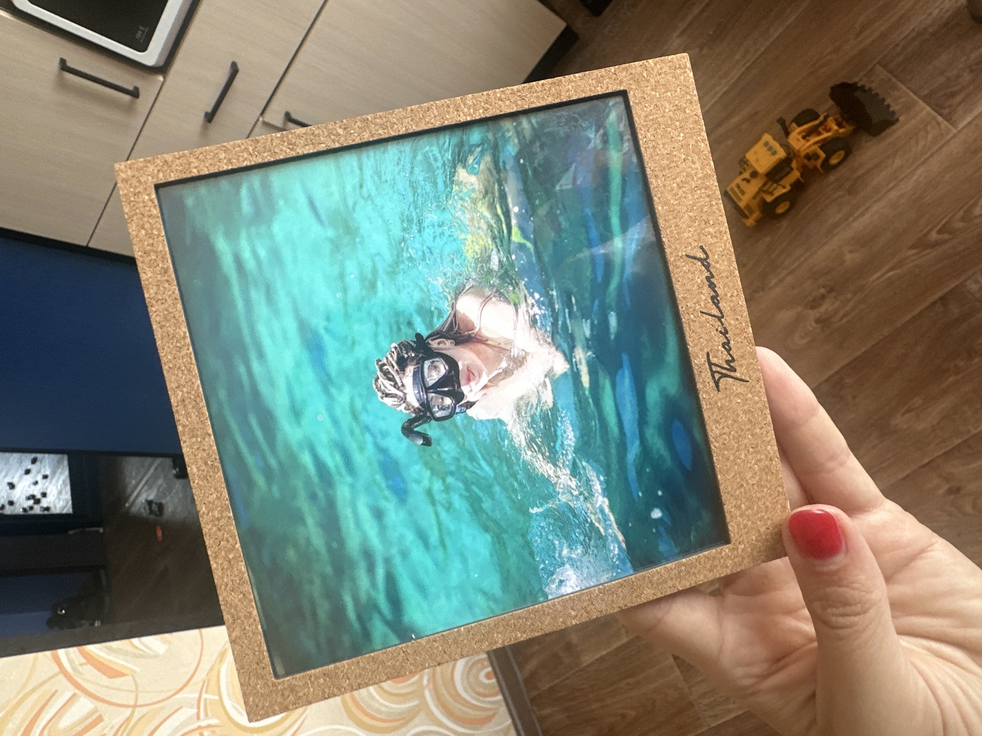 A framed photograph of a young woman snorkeling in turquoise water is held by a hand with red nail polish. The photograph, which has "Thailand" handwritten in script across the bottom, depicts the woman wearing a mask and snorkel. The frame itself is made of cork. The background of the image shows a wooden floor with a small yellow toy bulldozer placed on it, and part of a cabinet with handles is visible. The area appears to be indoors, potentially a home environment. No specific location within Novoaltaysk, Russia, can be identified from the image.