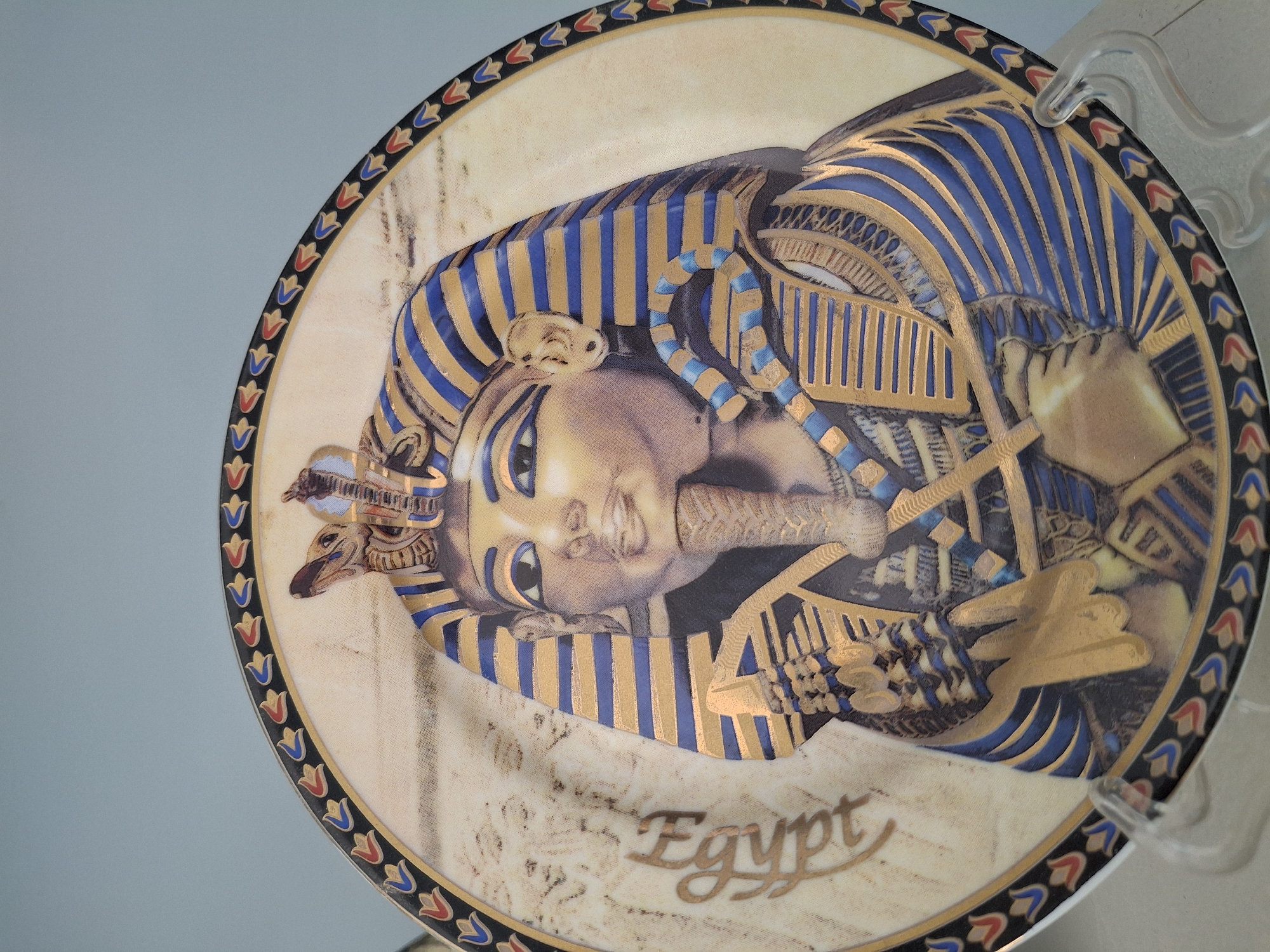 A decorative plate depicting an Egyptian pharaoh is mounted on a clear plastic stand against a light gray wall. The plate is round with a black border ornamented with small, stylized red and blue flowers. Within the border, the plate features a portrait of the pharaoh in traditional headdress with blue and gold stripes. The pharaoh's skin is a light tan color. The word "Egypt" is written in gold cursive lettering near the bottom of the plate. The plate's background has a mottled, light brown texture. There is no indication this image was taken in Pleven, Bulgaria.