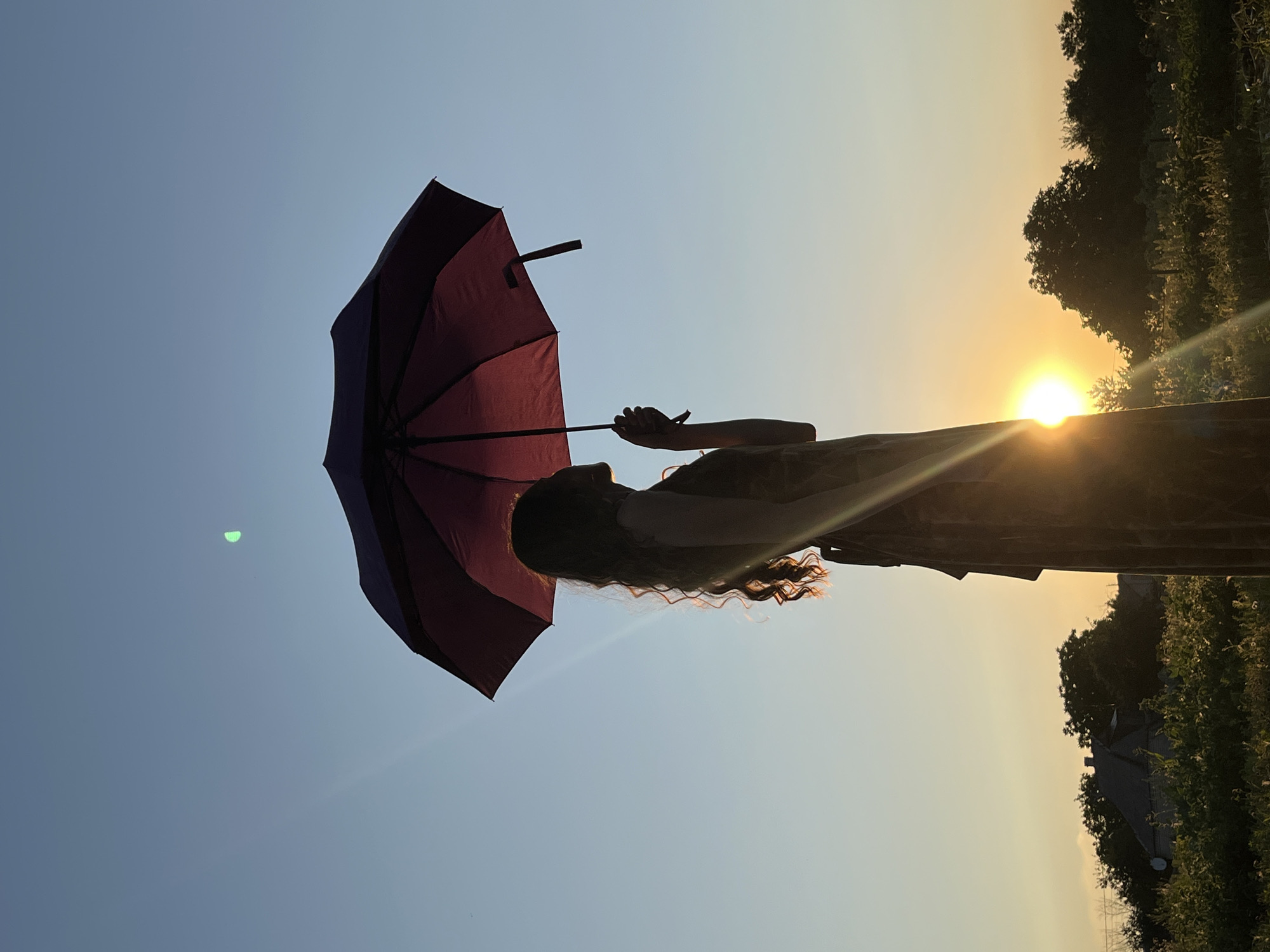 A silhouette of a young woman stands in profile, holding a closed, dark red umbrella above her head. She has long, wavy hair and is wearing a long, patterned dress. The woman is positioned against a clear blue sky with the sun setting in the background, casting a warm glow. A lens flare streaks diagonally across the image from the sun. In the distance, a treeline is visible with a house and some vegetation in the foreground. The overall scene depicts a serene outdoor setting at dusk.
