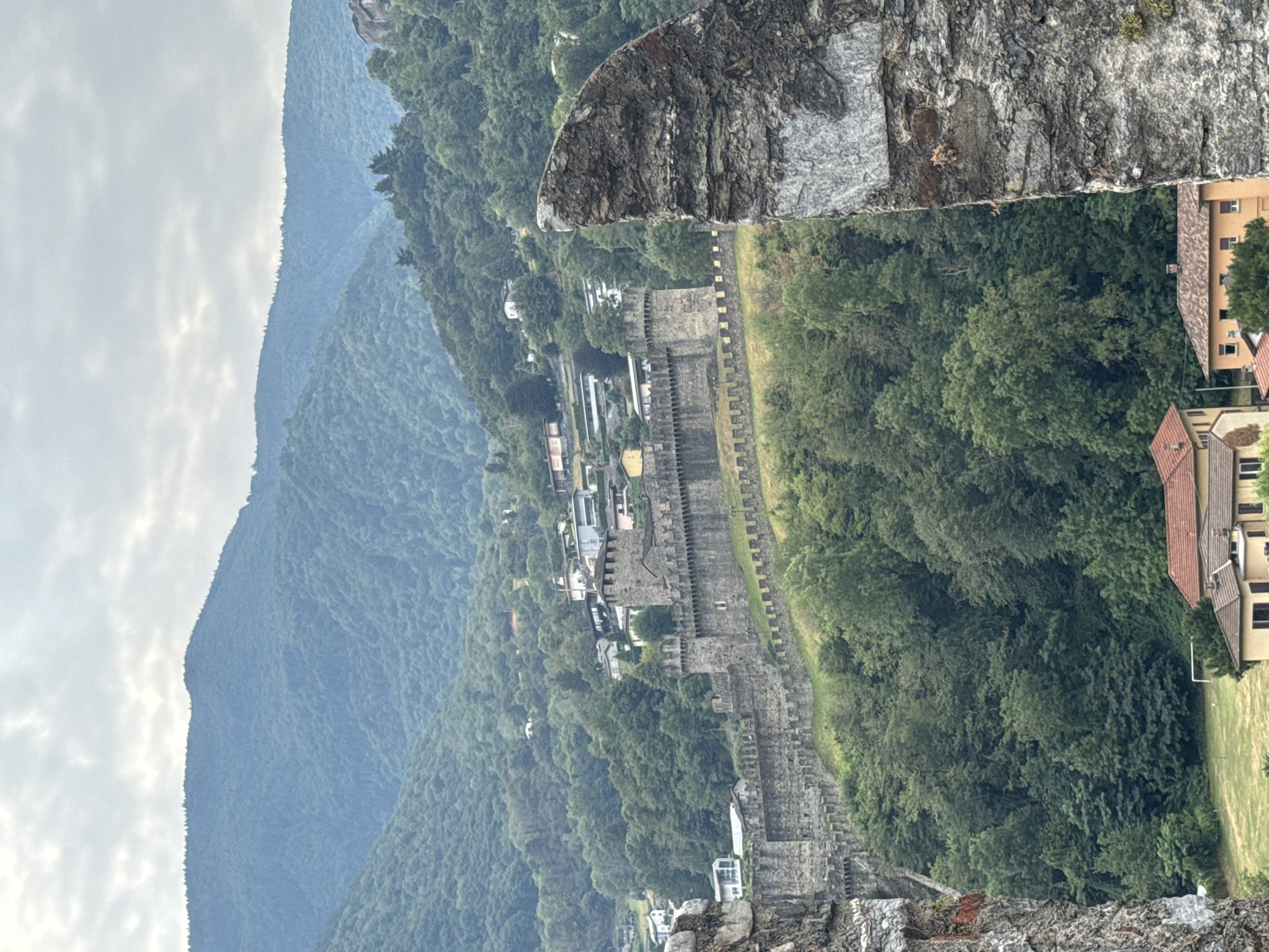 The image depicts a view of the Castel Grande in Bellinzona, Switzerland, framed by a rough stone foreground. The castle, a UNESCO World Heritage site, is a large medieval stone fortification situated on a hillside overlooking the city. It features crenellated walls and towers, with a section of the wall and a tower clearly visible. The castle is surrounded by lush green trees and a dense forest that extends up the mountainside in the background. A cluster of houses with red-tiled roofs is visible at the base of the hill, with more scattered houses higher up the slopes. The sky is overcast with soft, diffused light, suggesting a cloudy day. No people or animals are discernible in the image. The perspective suggests the photograph was taken from a higher vantage point, possibly another part of the castle complex or a nearby elevated structure.