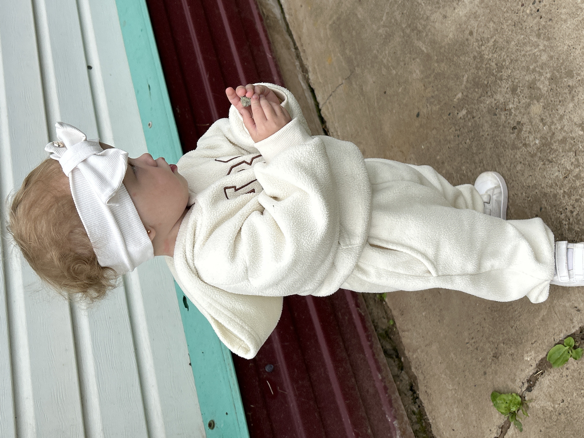 A young child, likely a toddler, is standing outdoors on a concrete surface. The child is wearing a cream-colored fleece sweatsuit and white sneakers. A white fabric headband with a bow is on the child's head. The child is holding a small, gray object in their hands, which they are examining. In the background, there is a building with horizontal siding painted in alternating sections of white and a dark reddish-brown, separated by a thin teal band. The ground consists of concrete paving stones with cracks and some sparse green vegetation growing through them. The scene appears to be in a residential area, possibly a backyard or a walkway. There is no direct indication of the city of Азинский, Russia, but the environment is consistent with a rural or suburban setting.