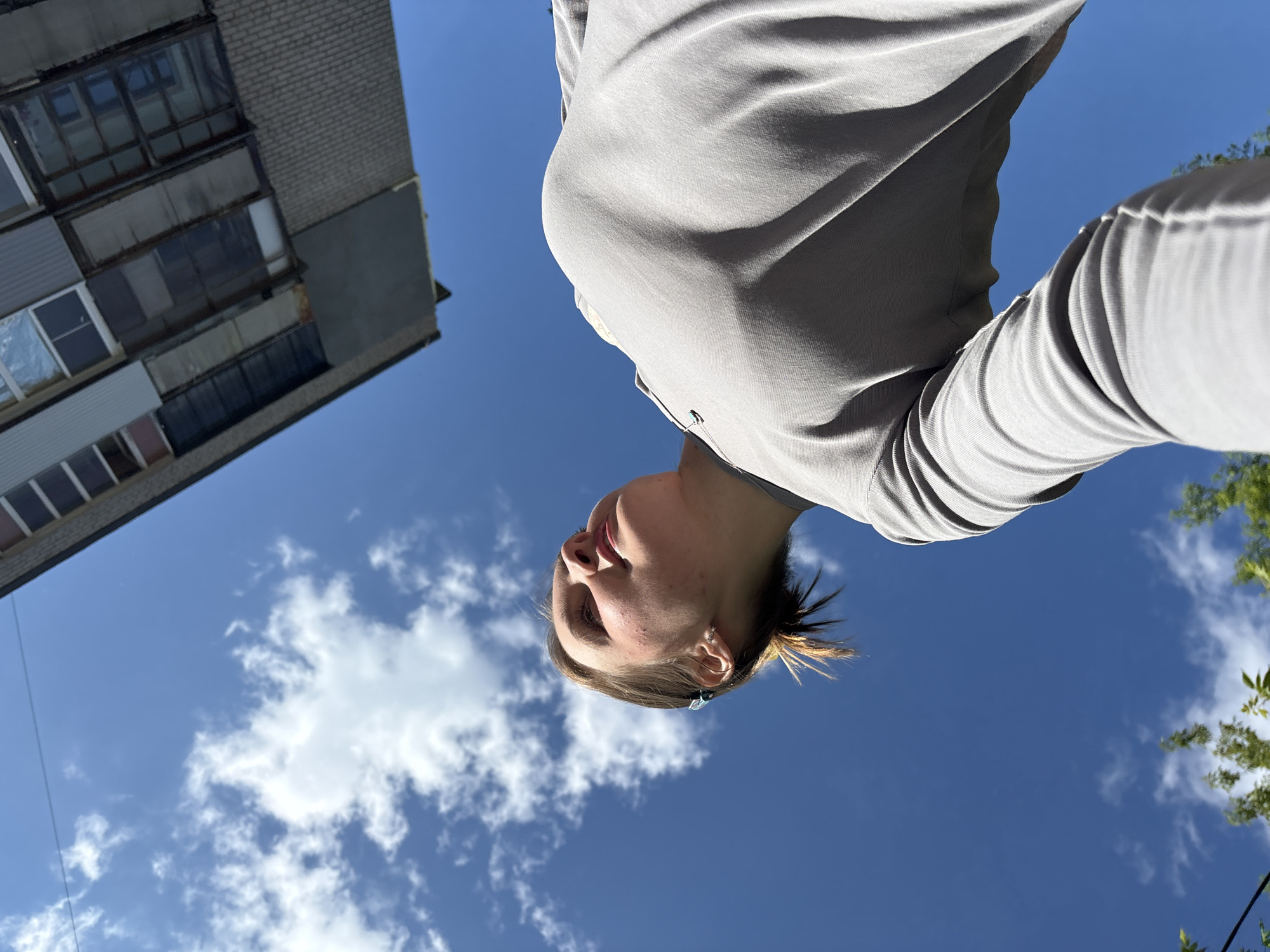 The image shows a low-angle view of a person with fair skin wearing a gray shirt, looking up at the sky. Their hair is light brown with blonde highlights, and they have a blue hair clip. The background consists of a blue sky with scattered white clouds, the top of a brick building with multiple windows on the left, and green trees on the right. The photo appears to be taken outdoors during daylight. While a specific location cannot be confirmed from this photo alone, the context suggests this image was possibly taken in Perm, Russia.