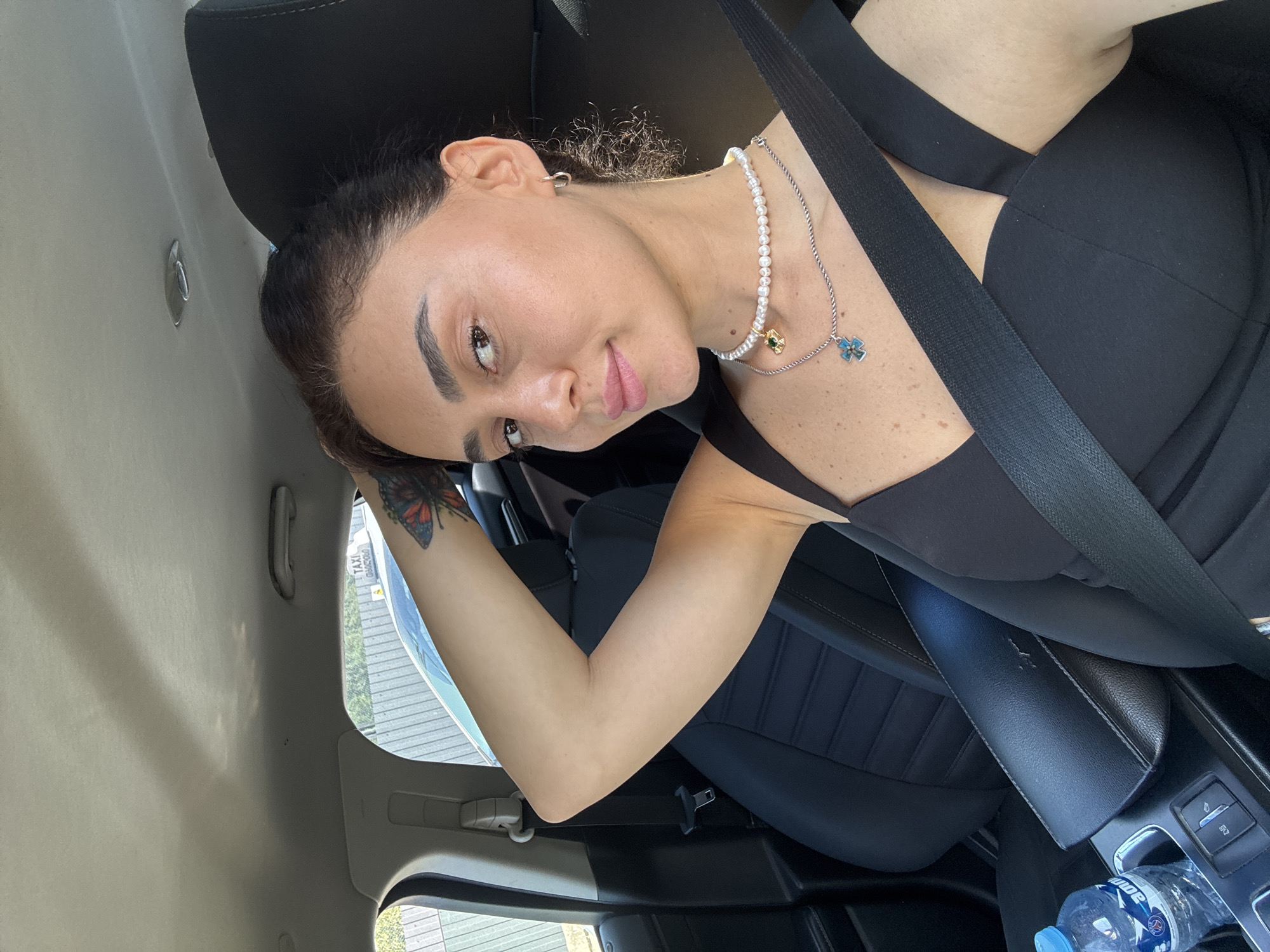 A woman with dark hair and light skin is sitting in the driver's seat of a car, with her left arm raised and hand resting on her head. She is wearing a black top and a seatbelt. The car's interior is visible, including the dashboard and a water bottle on the center console. Outside the car, a fence and a sign that reads "TAXI" can be seen. The scene appears to be in Tbilisi, Georgia.