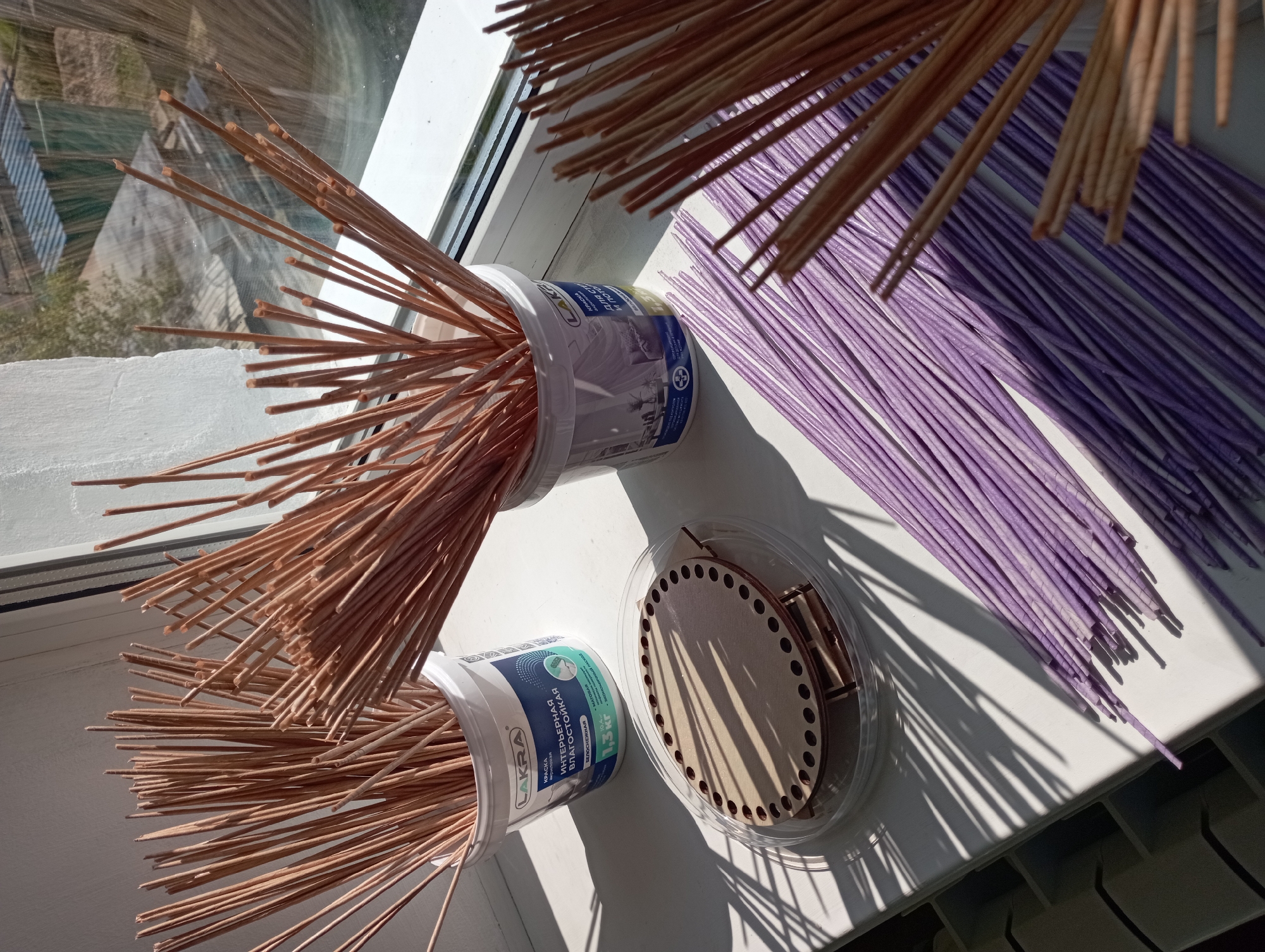 In Blagoveshchensk, Russia, on a sunlit windowsill, a collection of craft supplies is arranged. Two white plastic buckets, one labeled "LAKRA" with text in Russian indicating interior moisture-resistant paint, contain bundles of thin, brown wooden dowels. Beside them, a clear plastic container holds a circular wooden craft base with numerous pre-drilled holes and some smaller wooden pieces, possibly for assembly. Scattered in front of these are numerous thin, purple-colored dowels, also seemingly for crafting. The window behind offers a view of a slightly overgrown yard with corrugated metal roofing and trees. The sunlight casts strong shadows from the dowels onto the white surface of the windowsill.