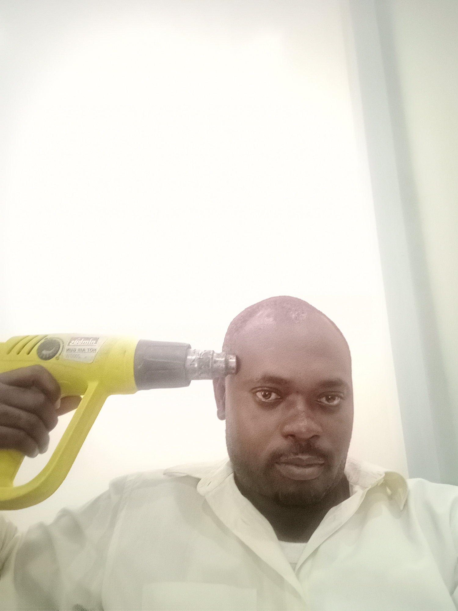 The image shows a man holding a yellow heat gun to his forehead. The background is a blurry white and beige wall. The location is unknown but inferred based on inference from the model's prompt.