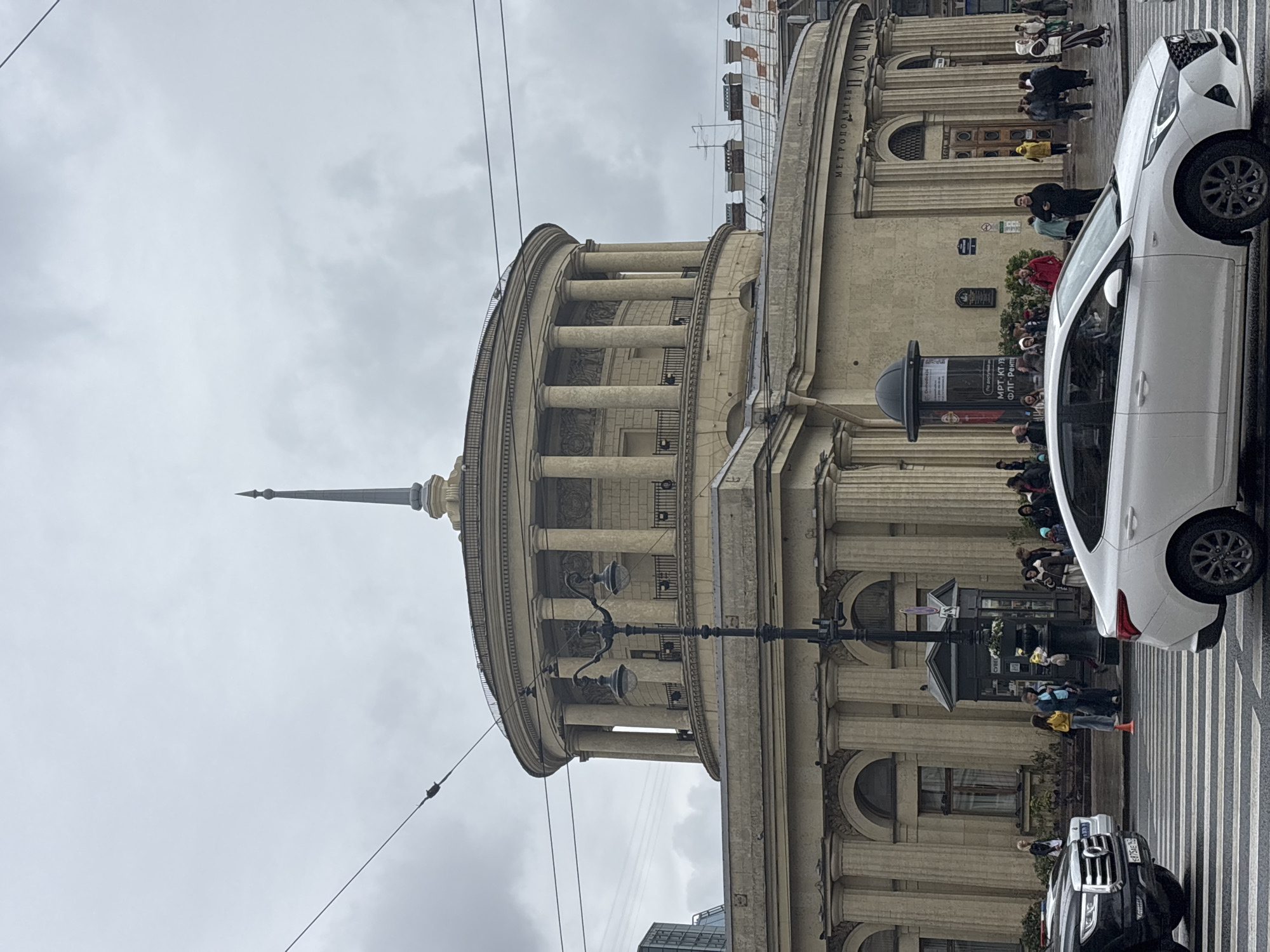 The image depicts a segment of the Moskovskaya metro station building in Saint Petersburg, Russia, taken from street level. The architectural style is neoclassical, with a rounded facade, columns, and a spire. A white car is parked near a crosswalk. Pedestrians are visible on the crosswalk and near the entrance of the metro station. A black service kiosk stands on the sidewalk. The sky is overcast.