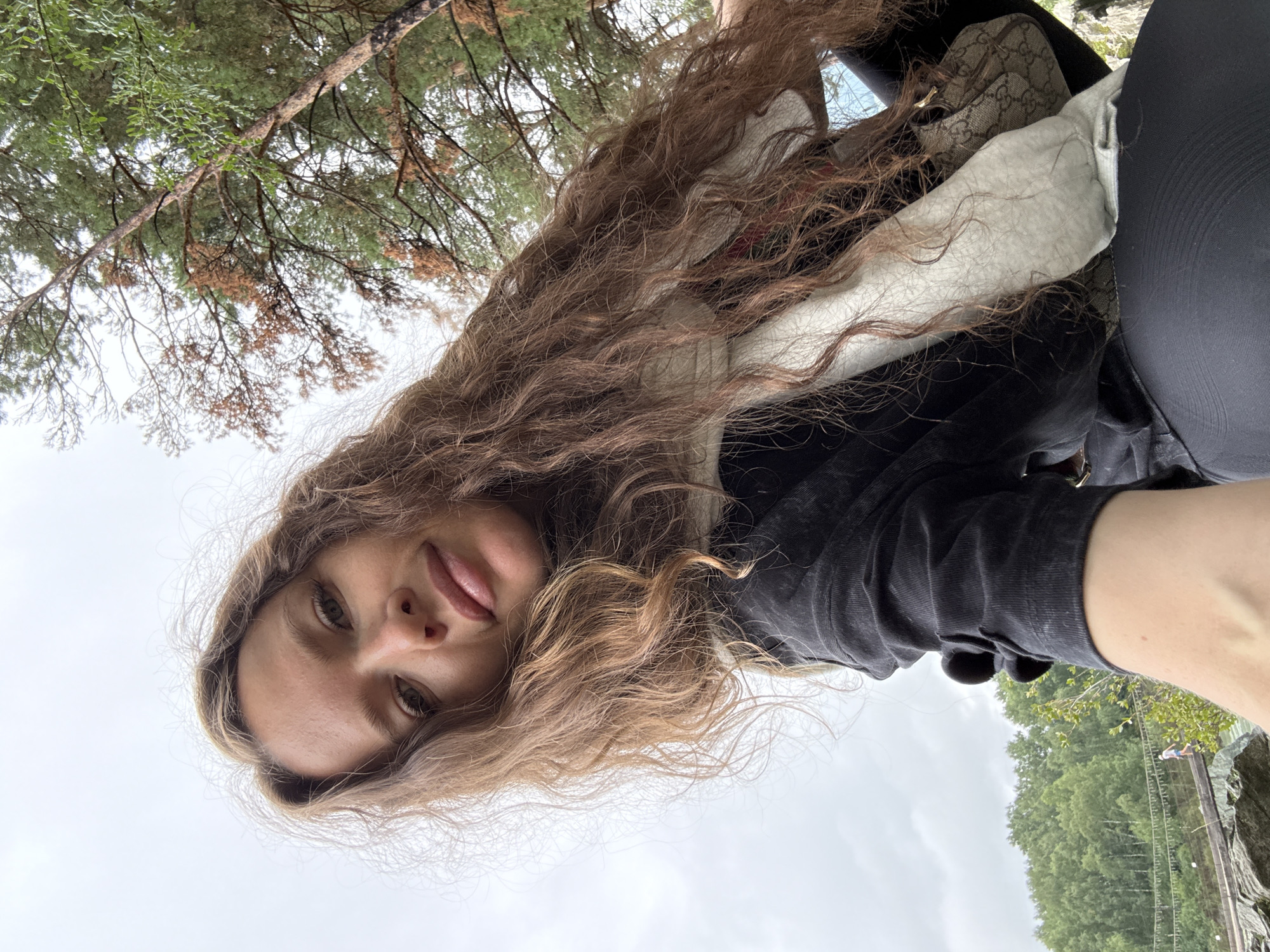 A woman with long, curly brown hair is taking a selfie outdoors. She is wearing a black jacket, a light-colored scarf, and dark leggings. A small Gucci bag is visible on her shoulder. In the background, there are trees and a cloudy sky. Further in the distance, there is a suspension bridge with a person walking across it, set against a backdrop of lush green trees and a body of water. This scene is consistent with the natural landscape found in Elekmonar, Russia, a region known for its scenic beauty and outdoor recreational opportunities, including suspension bridges like the one pictured.