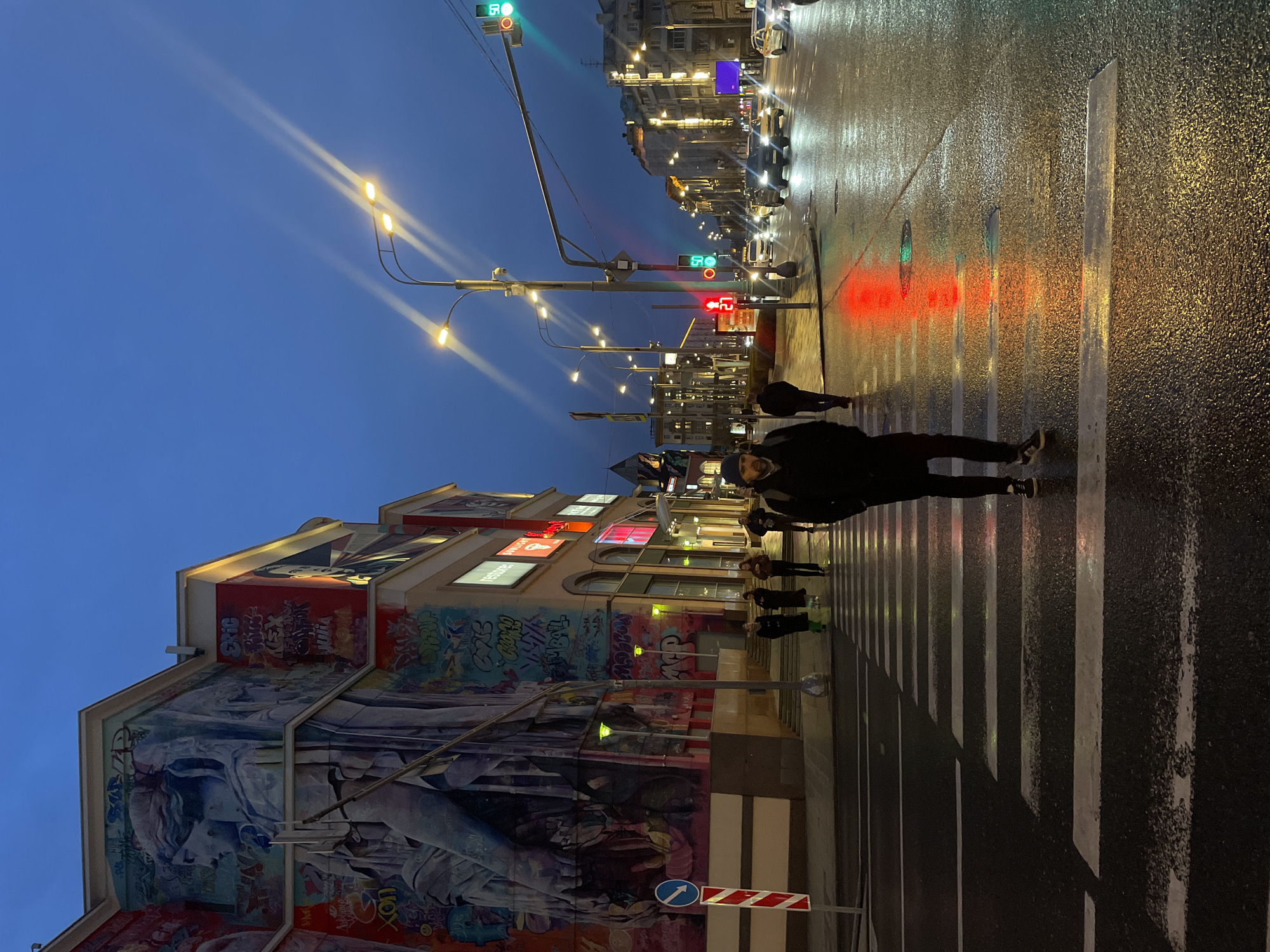 The image depicts a street scene in Moscow, Russia at dusk. The photograph is taken at street level at a crosswalk. The road surface is wet and reflects the city lights. A building with colorful graffiti covers most of one side of the street, and other buildings are visible further down the street. Several people are crossing the street in the crosswalk. Streetlights illuminate the scene and their light casts shadows. Traffic lights are visible; some are green and some are red. Cars can be seen in the distance.
