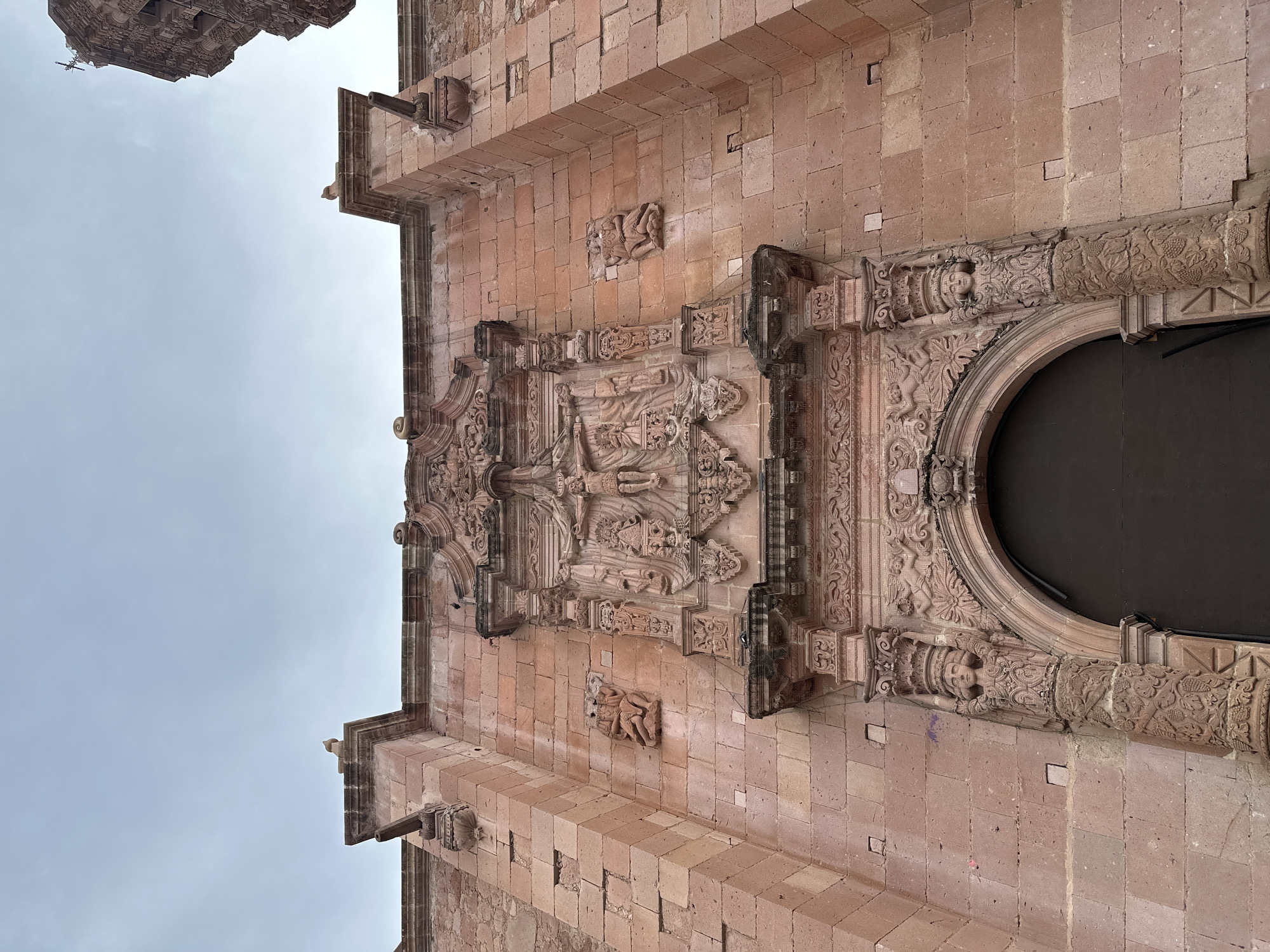 This is a low-angle exterior shot of a stone facade of a historic building, likely a church, in Zacatecas City, Mexico. The facade is constructed of large, pinkish-red sandstone blocks. The central focus is an ornate stone relief carving above an arched doorway. The relief depicts a crucifixion scene with Jesus on the cross, flanked by figures. Below this, smaller carvings include what appear to be cherubs or angels. Flanking the central relief are two pilasters, each topped with a carved figure supporting the elaborate entablature. The pilasters themselves are decorated with intricate patterns, including what seems to be a vine motif. On the left and right sides of the main facade, higher up, are two small, figural carvings in relief, appearing to depict seated figures. Above the facade, the sky is overcast with a pale blue hue. The overall impression is of weathered, detailed stonework typical of colonial architecture.