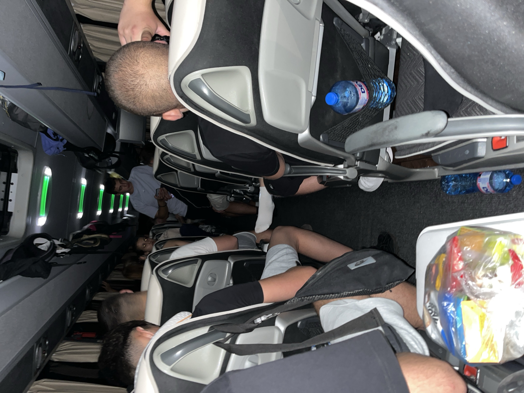 Inside a bus, passengers are seated in rows. Aisle lighting emits a green glow. Overhead compartments line the ceiling. Two blue water bottles are visible in seat back pockets. A bag of snacks rests on a tray table. The location is Dragoman, Bulgaria.