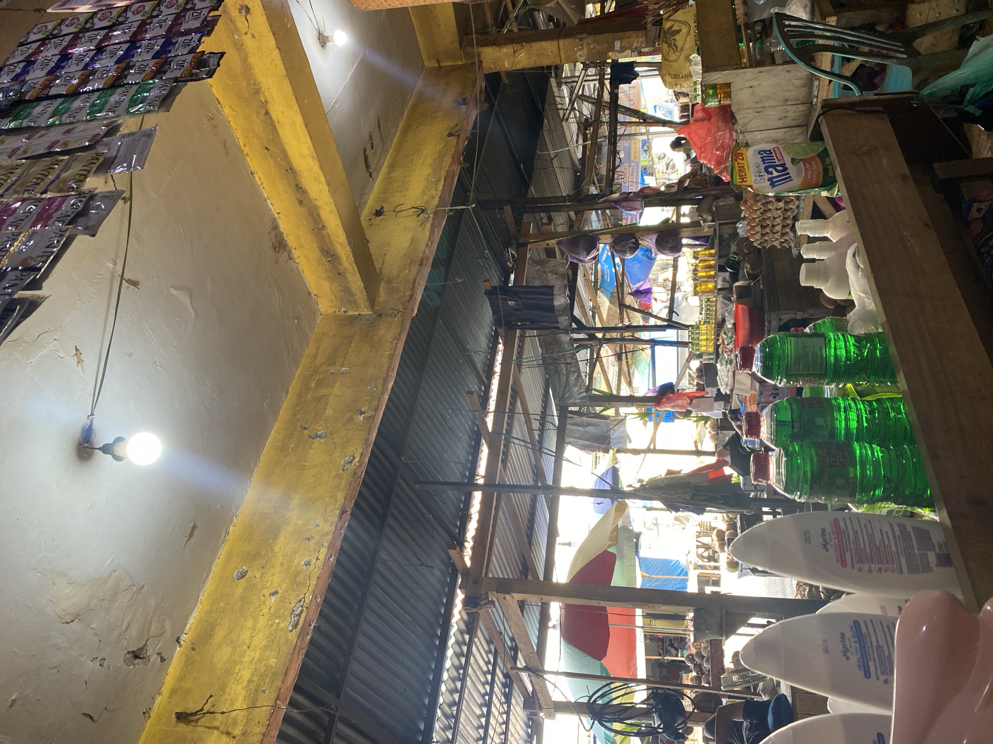 The image depicts an indoor market stall, likely in Ta', Indonesia, characterized by a rustic, somewhat dilapidated setting. A bright yellow, distressed beam runs across the top of the frame, with a corrugated metal roof visible above it. The ceiling itself is a pale, textured surface with cracks and water damage, illuminated by a single bare light bulb.
Below the yellow beam, the market unfolds. Numerous stalls are visible, constructed from wood and corrugated metal, creating a layered, sheltered environment. Colorful umbrellas – red, green, and yellow – are set up, suggesting outdoor sections or temporary coverings. Various goods are displayed: rows of small plastic packets, possibly shampoo or seasoning, hang from the ceiling in one area. Elsewhere, bottles of green liquid, likely cleaning products or beverages, are lined up. Further back, stacked boxes of eggs and what appear to be coconuts are visible. A red plastic bag and other assorted items are scattered on shelves.
In the foreground, several white plastic bottles with curved tops are prominently displayed on a wooden counter. To their right, three green plastic bottles are arranged. A partial view of a pink object is visible on the left.
Several people are present in the scene, though mostly indistinct. A person is seated in the background, partially obscured. Another individual, seen from the back, is wearing a dark top and appears to be interacting with the stall. A person in a light-colored top is also visible further in the background. A light blue chair is positioned to the right of the frame, near a pile of sacks. The lighting is uneven, with strong sunlight filtering in from the back and the artificial light from the bulb creating shadows and highlighting the textures of the environment.