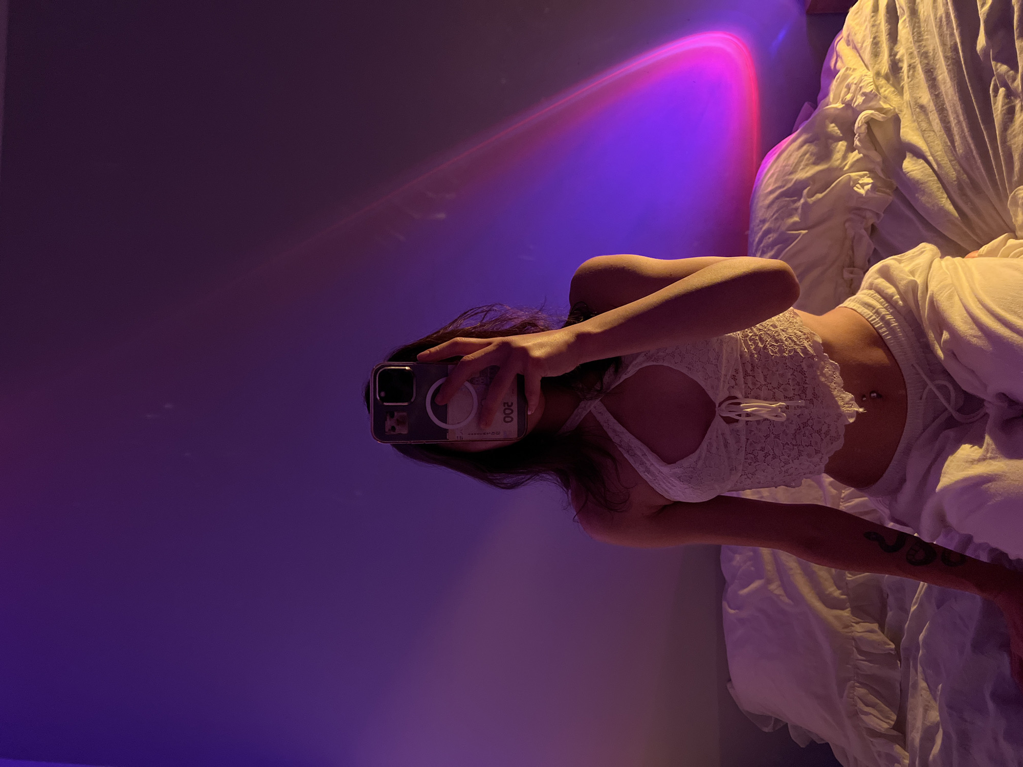 A person is taking a selfie in a bedroom. The person is sitting on a bed with white bedding. They are wearing a white lace top and white pants. The person is holding a phone in front of their face, partially obscuring their face. A tattoo of a snake is visible on their left arm. The room is dimly lit, with purple and pink light casting a glow on the wall and bed. The wall has a projected light effect that creates a gradient of colors. There are white pillows with ruffled edges on the bed. The scene is set in a bedroom. The image does not contain any information that can be used to determine the location in the city of Ternopil, Ukraine.