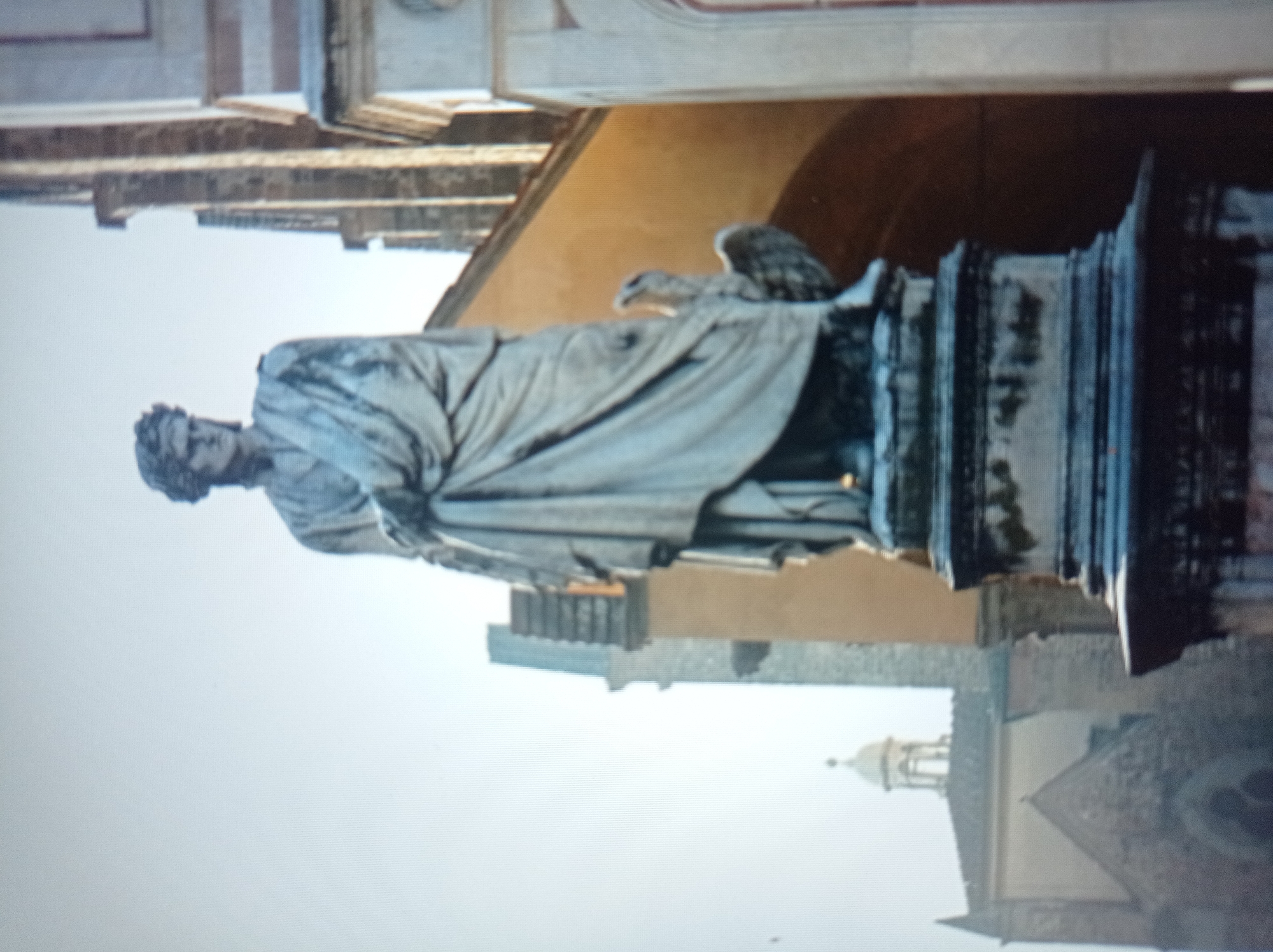 The image shows a full-length white stone statue of a robed man wearing a laurel wreath on his head. He stands on a pedestal. To his right, a stone eagle with spread wings is depicted, also on the pedestal. In the background, there are buildings with arched windows and towers. The statue is centrally located in the frame. This statue is located in the city of Mukachevo, Ukraine.