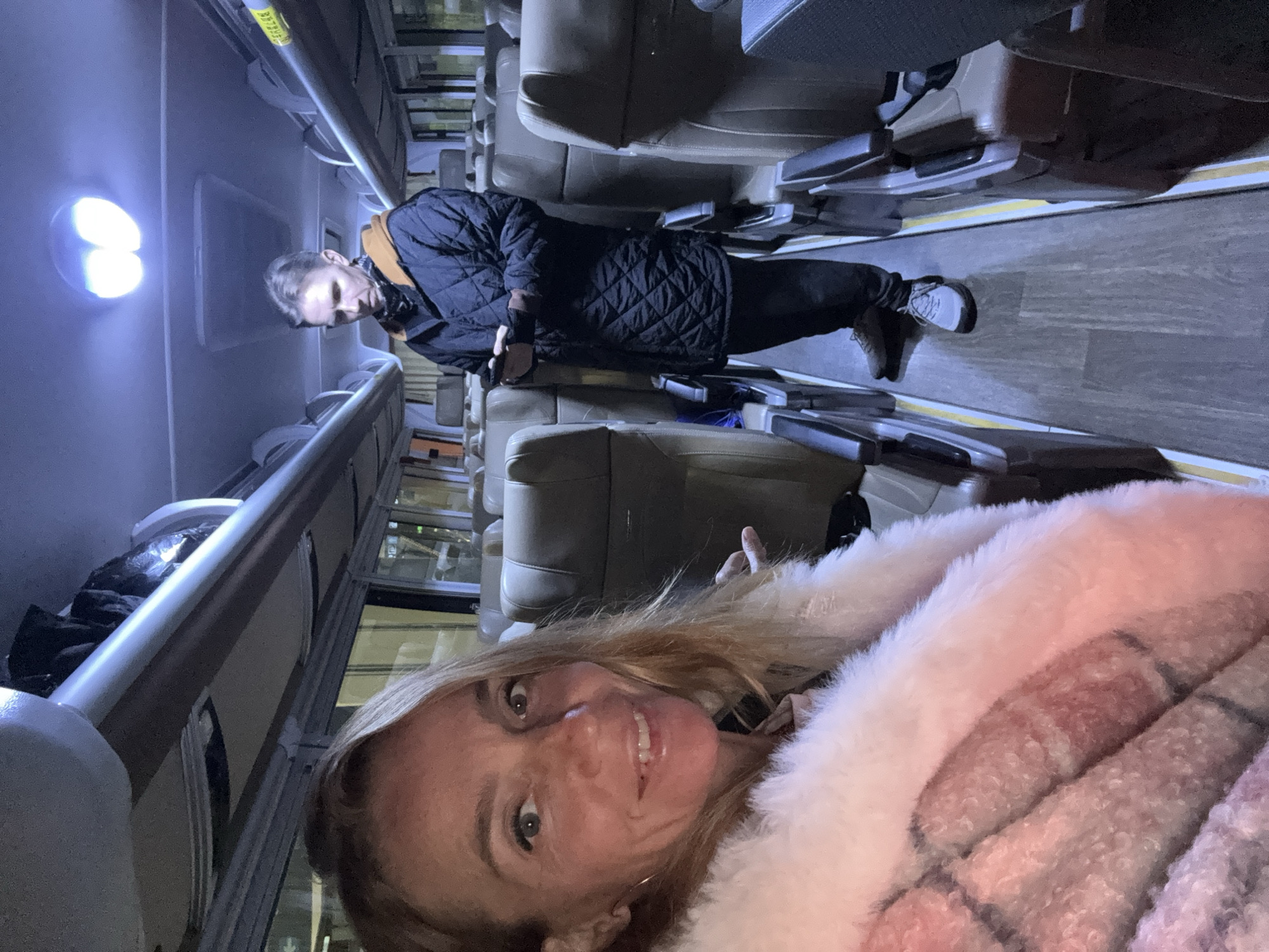 The image appears to be taken inside a bus or train carriage, illuminated by overhead lighting. A woman is taking a selfie in the foreground, partially obscured by a pink and white furry coat. In the background, a man is standing, wearing a black jacket and dark pants, looking down at something in his hands. Rows of tan-colored seats line the interior of the vehicle. Based on the presence of the bus and the implied location in Wulong District, China, it is likely public transport is shown.