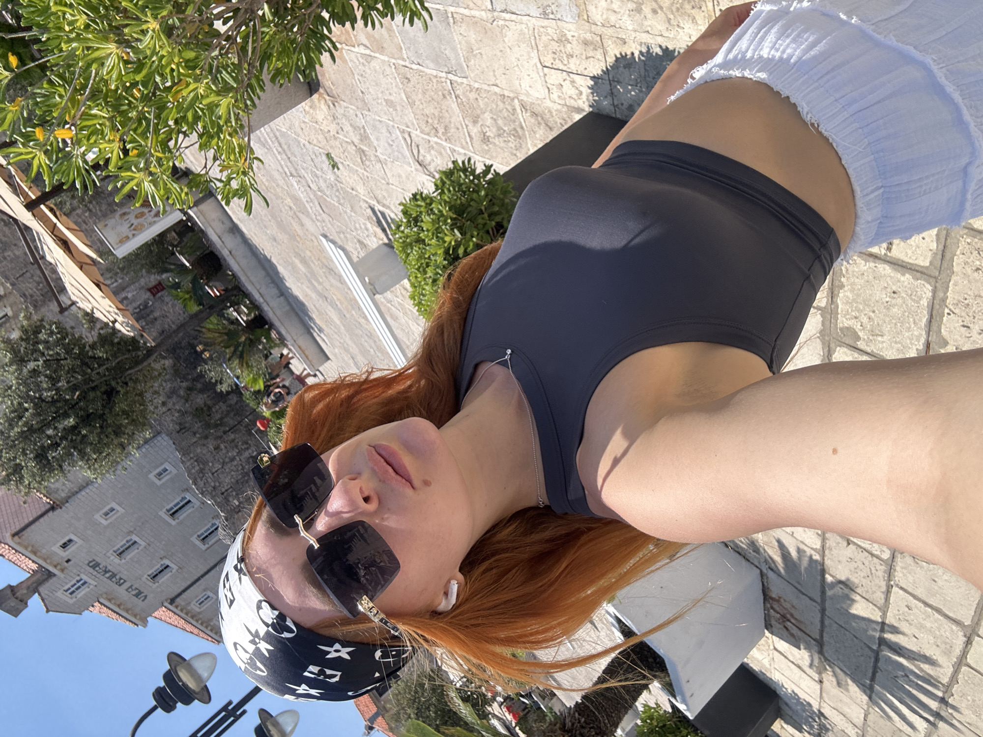 A woman with long, reddish-blonde hair takes a selfie outdoors. She is wearing a black bandana with a white pattern, large black sunglasses, a dark grey crop top, and a white ruffled skirt. She has white earbuds in her ears. She is standing on a paved area with sunlight casting shadows from palm fronds. In the background, a stone building with the words "VILA BUKOVAC" is visible, along with some greenery and a streetlamp. The scene appears to be in Budva, Montenegro.