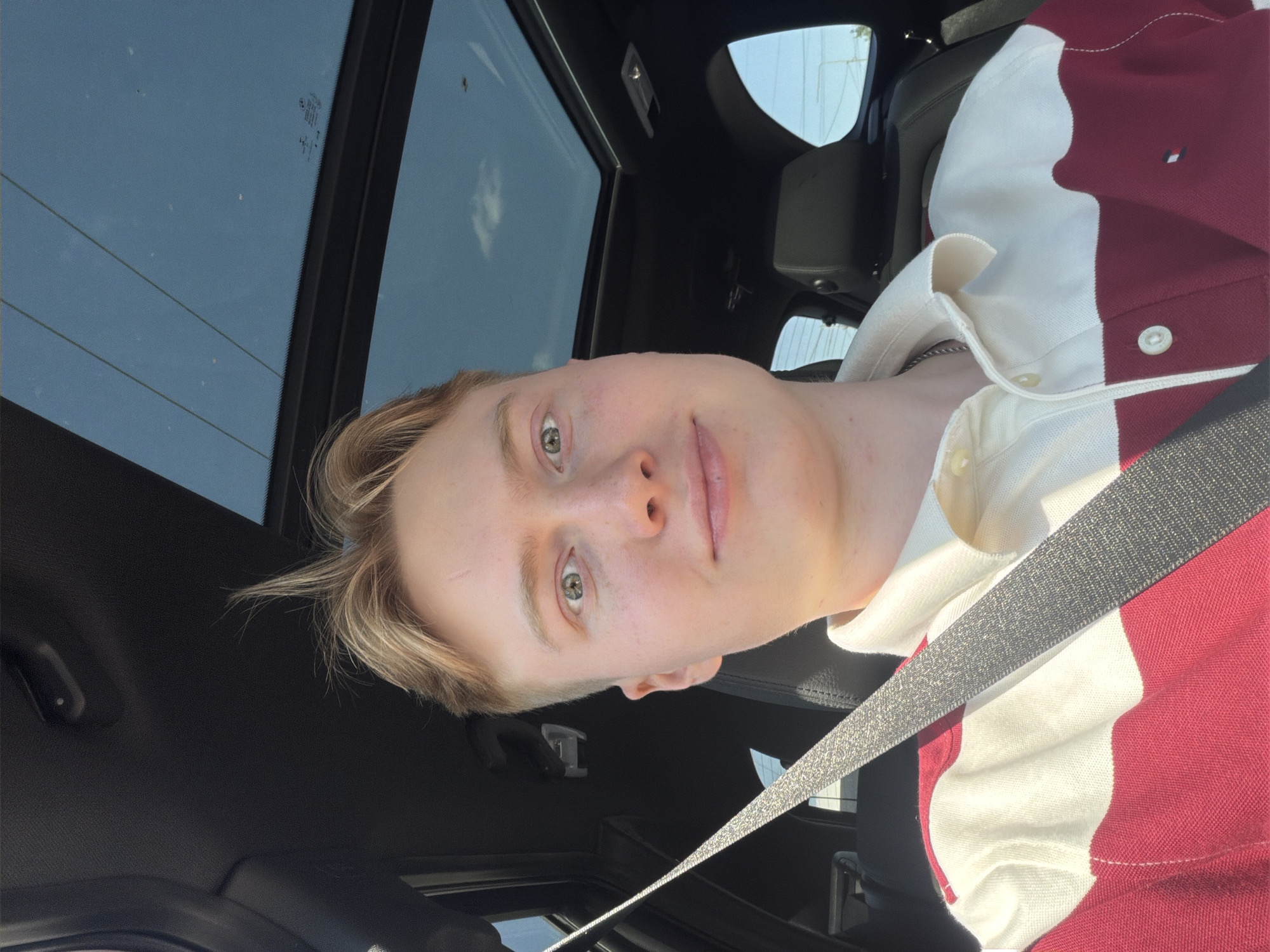 A young man with blond hair is seated in the driver's seat of a car, looking towards the camera. He is wearing a collared shirt with white and burgundy stripes. The car has a sunroof, which is open, revealing a clear blue sky. The interior of the car appears to be black leather. A seatbelt is visible diagonally across the man's chest. There are no discernible landmarks or specific features that would allow for the identification of the location as Kyiv, Ukraine. The scene is a casual, everyday moment captured from inside a vehicle.