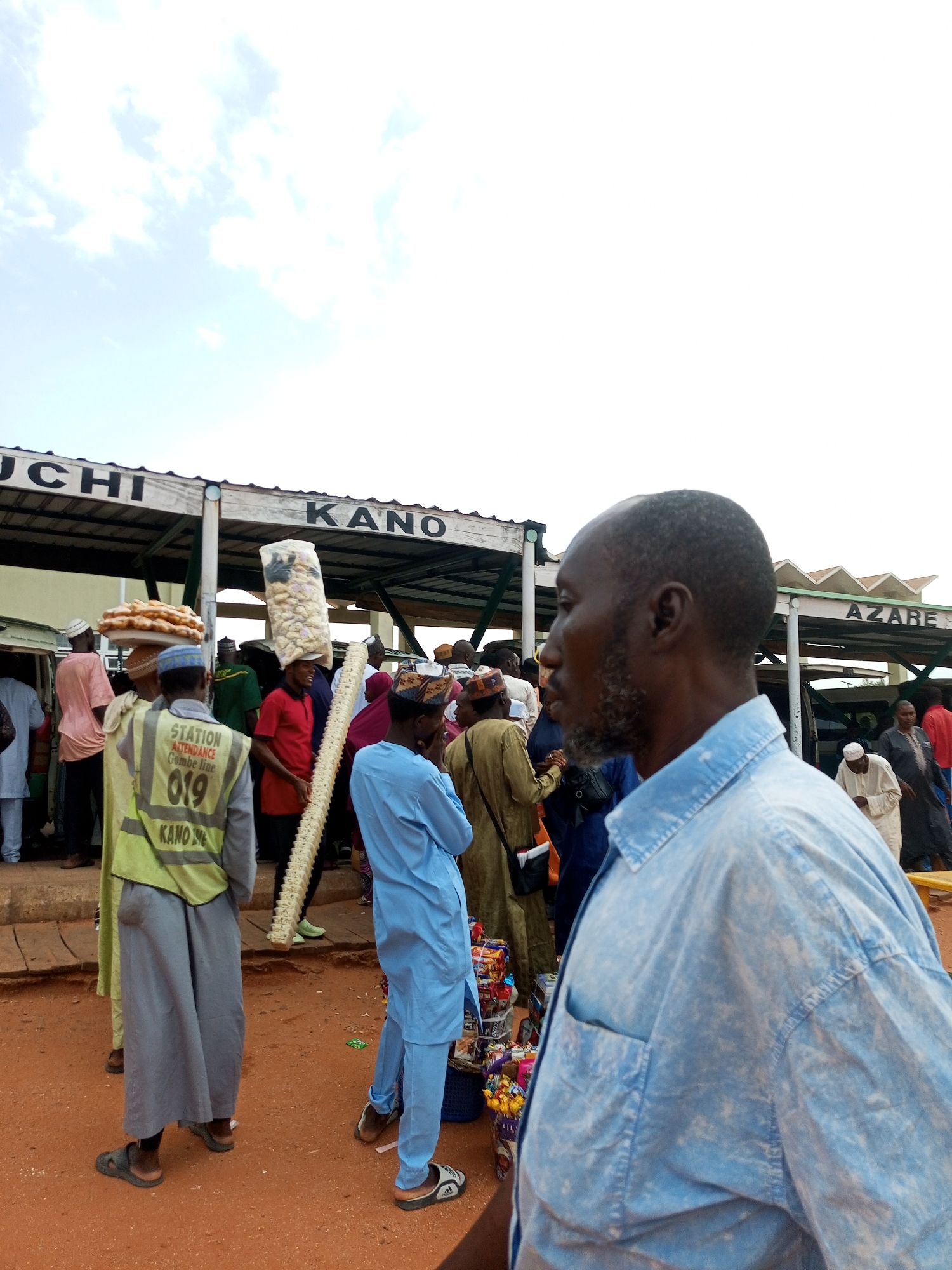 A man in a light blue denim shirt is in the foreground, facing away from the camera and to the left. He has a gray beard and a bald head. In the background, there are several people in traditional clothing, some carrying goods. A sign above them reads "UCHI KANO". Another sign in the background says "AZARE". The ground is covered in reddish-brown dirt. The scene appears to be a marketplace or transportation hub.