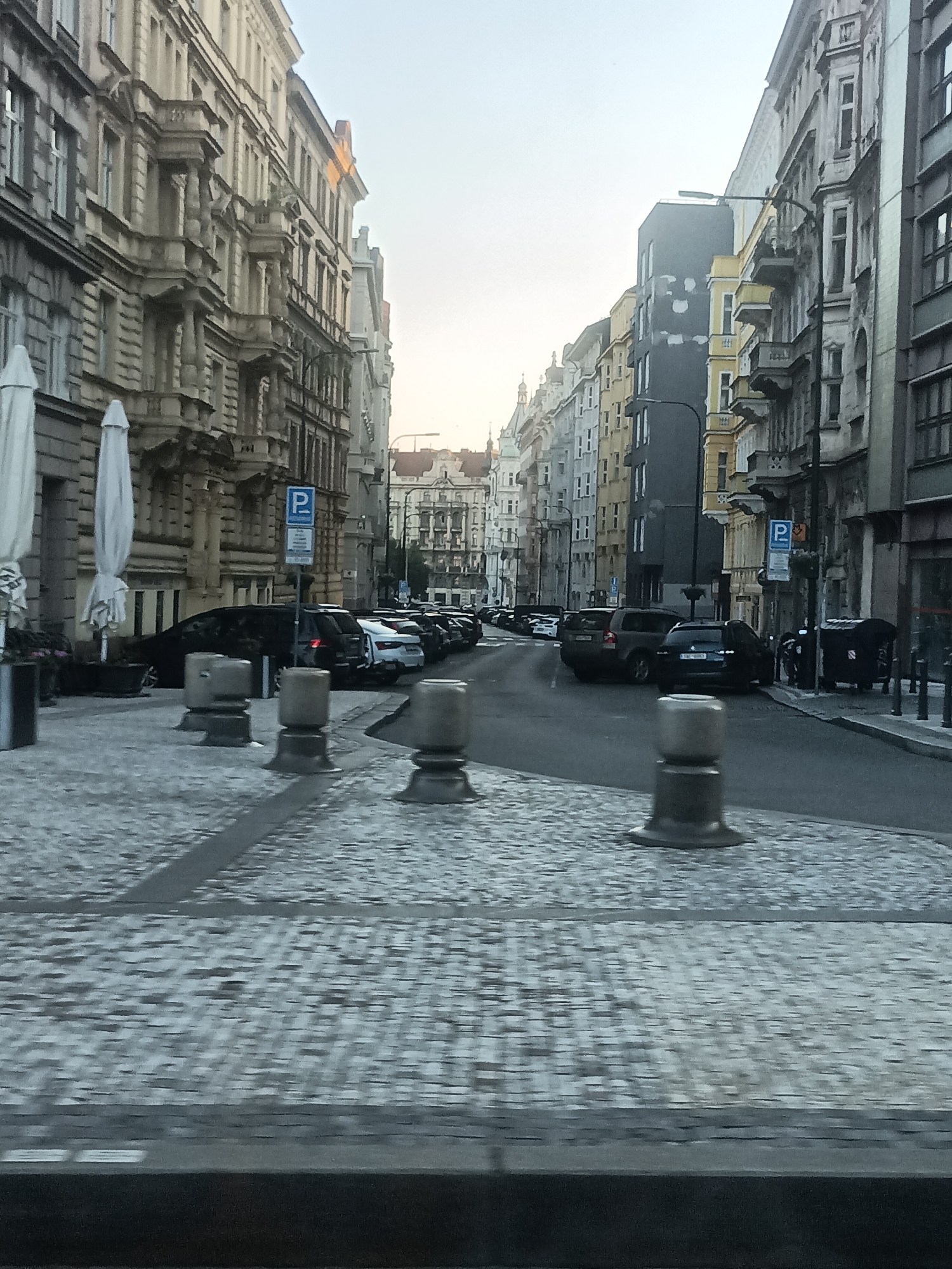 The image shows a street scene in Prague, Czechia, with ornate historic buildings lining both sides of a cobblestone street. Parked cars are visible along the right side of the street. In the foreground, there are several concrete bollards. The sky is overcast, and the overall lighting suggests it is either early morning or late afternoon. There are no people clearly visible in the image.
