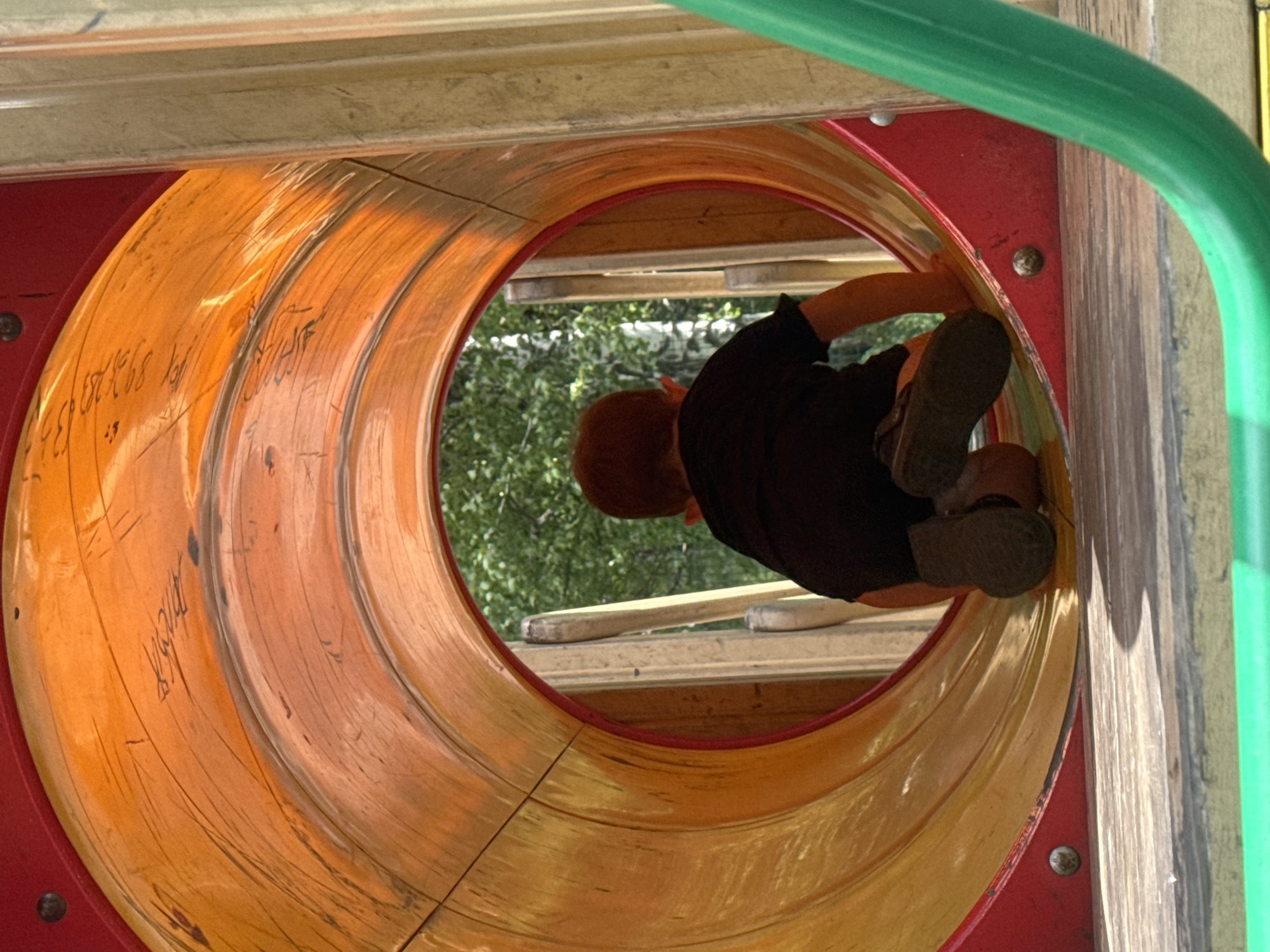 The image shows a young child crawling through a tubular playground slide. The child is wearing a black shirt and dark shoes. The slide is made of brightly colored plastic with orange and yellow sections, and it appears to be outdoors. The inside of the slide has some scuff marks and what looks like faint graffiti. Sunlight is filtering through the trees visible outside the slide, suggesting it is daytime with fair weather. The child's posture indicates they are actively moving through the tunnel. The setting appears to be a playground. No visible text is clearly legible.