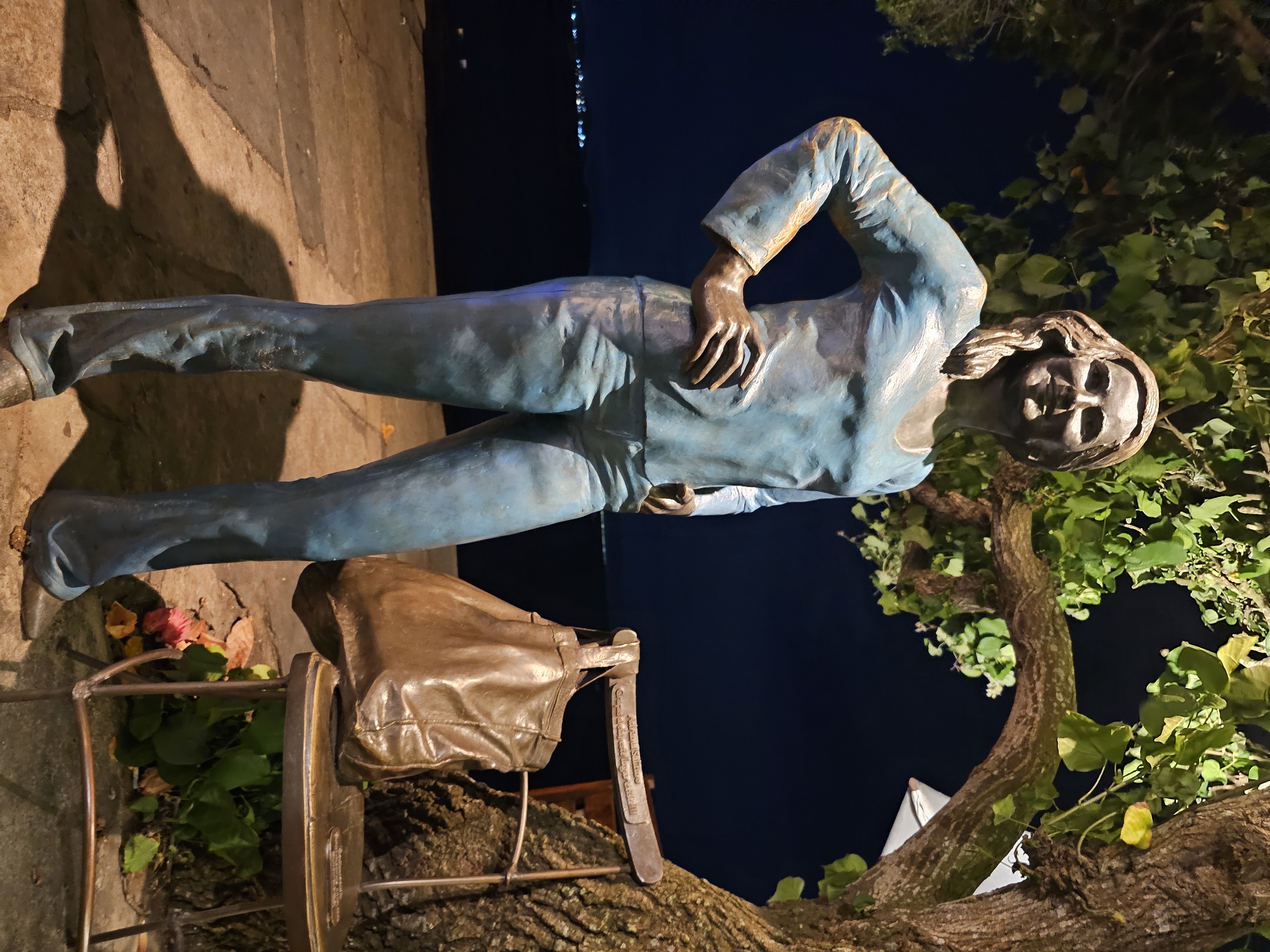Here is a description of the image:
This image shows a bronze statue of Brigitte Bardot reclining in Armação dos Búzios, Brazil. Bardot is in a blue suit, casually reclined against the background of the night, with a leather bag, and a leafy tree. In the dark distance there are lights on the water, and the foreground is a lit-up patio area.