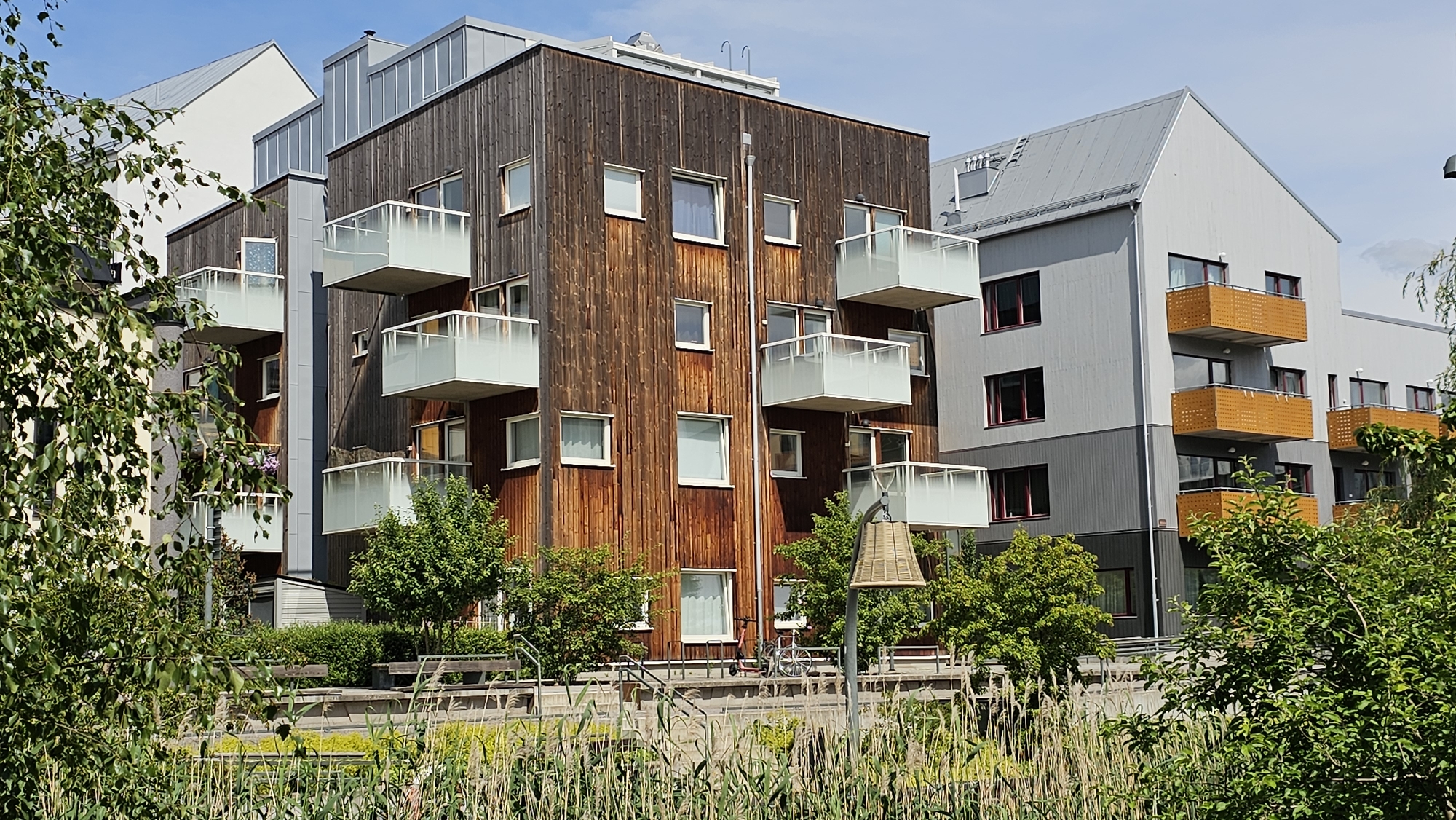 This media file depicts a modern apartment building complex with a blend of natural and man-made elements. The primary subjects are the multi-story buildings, characterized by their contemporary architectural design. One building features a facade of vertical wooden planks, complemented by white-framed windows and protruding balconies with glass railings. Another adjacent building has a lighter gray exterior, with recessed balconies featuring wooden railings and dark-framed windows. A third, taller building, partially visible on the far left, has a white facade and a distinctive angular roof.
The setting appears to be a residential area, with lush greenery in the foreground and surrounding the buildings. Tall reeds and grass grow along a body of water or wet area in front of the structures, suggesting a park-like or natural landscape. Trees and bushes are interspersed around the buildings, contributing to a serene and possibly urban-nature integrated environment.
There are several objects visible, including benches and stairs leading towards the buildings, implying public access or communal areas. A bicycle and an electric scooter are parked near the wooden-clad building, indicating the presence of residents or visitors engaging in recreational activities or transportation. A unique bell-shaped light fixture with a woven lampshade stands in the middle ground.
The scene is set under a clear blue sky with some wispy clouds, suggesting fair weather. The lighting indicates it is daytime, likely late morning or afternoon, given the presence of shadows and the brightness of the scene. No people are directly visible, and the overall mood conveyed is one of peaceful, modern living within a green environment. There is no visible text in the image. The location context is Linköping, Sweden, which aligns with the architectural style and the presence of ample green space.